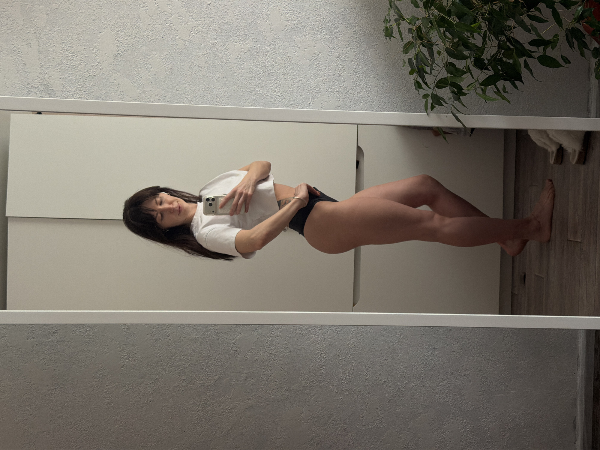 A young woman is captured taking a mirror selfie in an indoor setting, likely in Perm, Russia. The image shows her full-body reflection in a large, vertically oriented mirror. She has long, dark hair with bangs and appears to be looking at her smartphone, which she holds up to capture her image. Her expression is neutral, with her eyes slightly closed or downcast.
She is wearing a simple white crop top that exposes her midriff and dark, high-cut briefs or underwear. A bracelet is visible on her left wrist, and a tattoo can be partially seen on her left rib cage area. Her bare feet are visible, and she appears to have a fit physique.
The background reflected in the mirror consists of minimalist white cupboards or wardrobe doors, suggesting a clean and modern room aesthetic. To the woman's left (right side of the reflected image), a textured grey wall is visible. To her right (left side of the reflected image), a lush green plant with numerous leaves hangs down, adding a touch of nature to the indoor space. The room also features wooden flooring. The lighting appears to be bright and even, likely from indoor sources, illuminating the scene clearly. No discernible text is visible in the image.