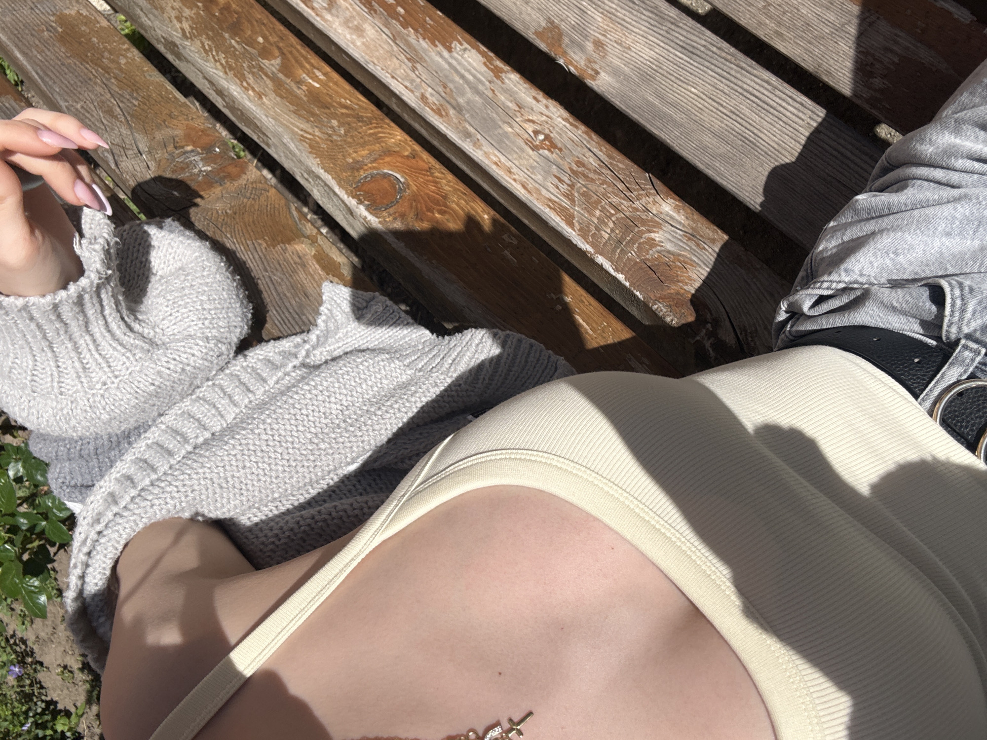 A high-angle shot captures a section of a person sitting on a weathered wooden park bench in what is likely Sofia, Bulgaria, although the city's specific location cannot be determined from the image alone. The person is wearing a light-colored tank top, a grey knitted sweater, and grey jeans with a black belt visible. A necklace with small pendants is visible on the person's chest. The bench's aged wood shows wear and tear, with sunlight casting shadows across its surface. Some greenery appears in the bottom left corner of the frame. The person's left hand, sporting long, manicured nails, is visible in the upper left corner.