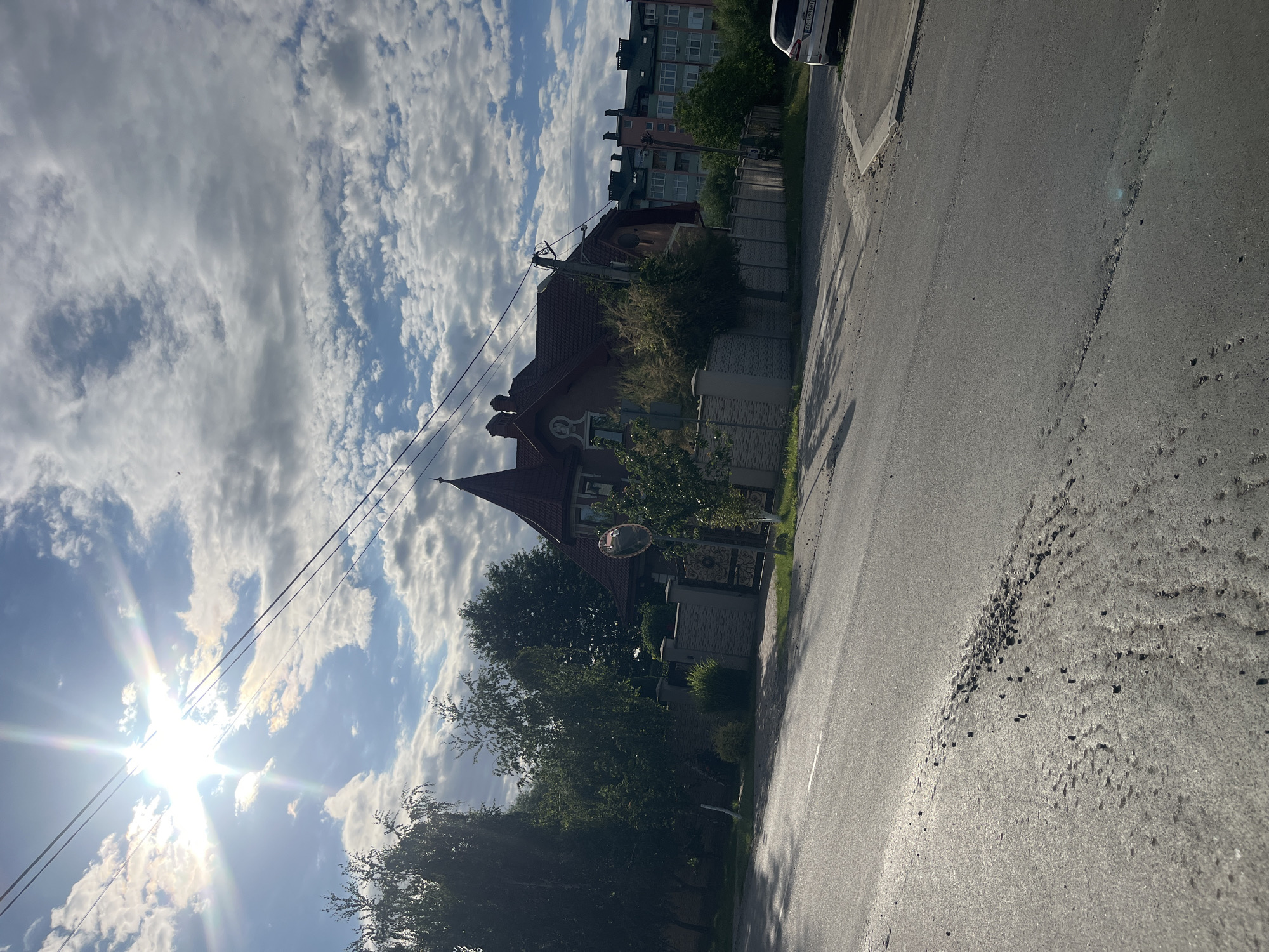 The image depicts a street scene in Storozhnytsia, Ukraine, under a bright, partly cloudy sky. The sun is prominently visible in the upper left quadrant, casting lens flares and a strong glare across the sky and the scene. The foreground is dominated by an asphalt road, showing some wear and tear with gravel scattered across its surface in a diagonal pattern. To the right, a white car with license plate AO 3793 HH is parked.
Behind the road and to the left, a residential property is visible. It features a light-colored fence with a decorative gate. Behind the fence, a house with a distinctive dark red, multi-faceted roof and an imposing spire-like turret is partially obscured by lush green trees. Further in the background, beyond the property, a multi-story apartment building with a green roof and light-colored facades can be seen. The overall impression is a suburban or semi-urban environment with a mix of residential buildings and green spaces. Power lines are strung across the upper portion of the image. No people are visible in the frame.