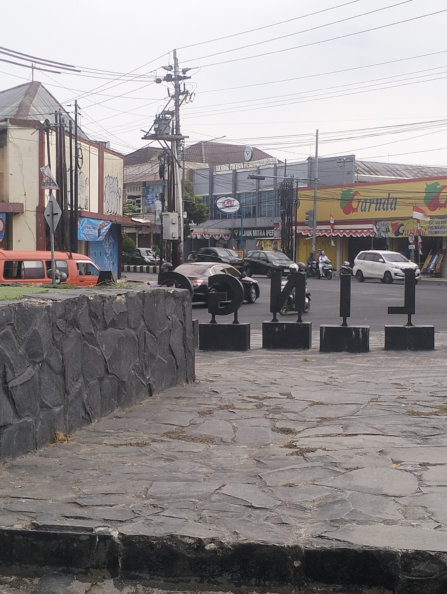 The image shows a street scene in Purbalingga, Indonesia, with a focus on an intersection and surrounding buildings. In the foreground, a stone wall and paving stones are visible. Across the street, several vehicles are present, including an orange van, a black sedan, and a white SUV. Motorcycles with riders are also seen navigating the intersection. Buildings lining the street include a structure with a red and yellow sign that reads "Garuda," suggesting a commercial establishment. Other buildings appear to be shops or offices. Overhead, utility poles with numerous wires crisscross the sky, indicating urban infrastructure. The overall atmosphere suggests a typical day in a bustling Indonesian city.