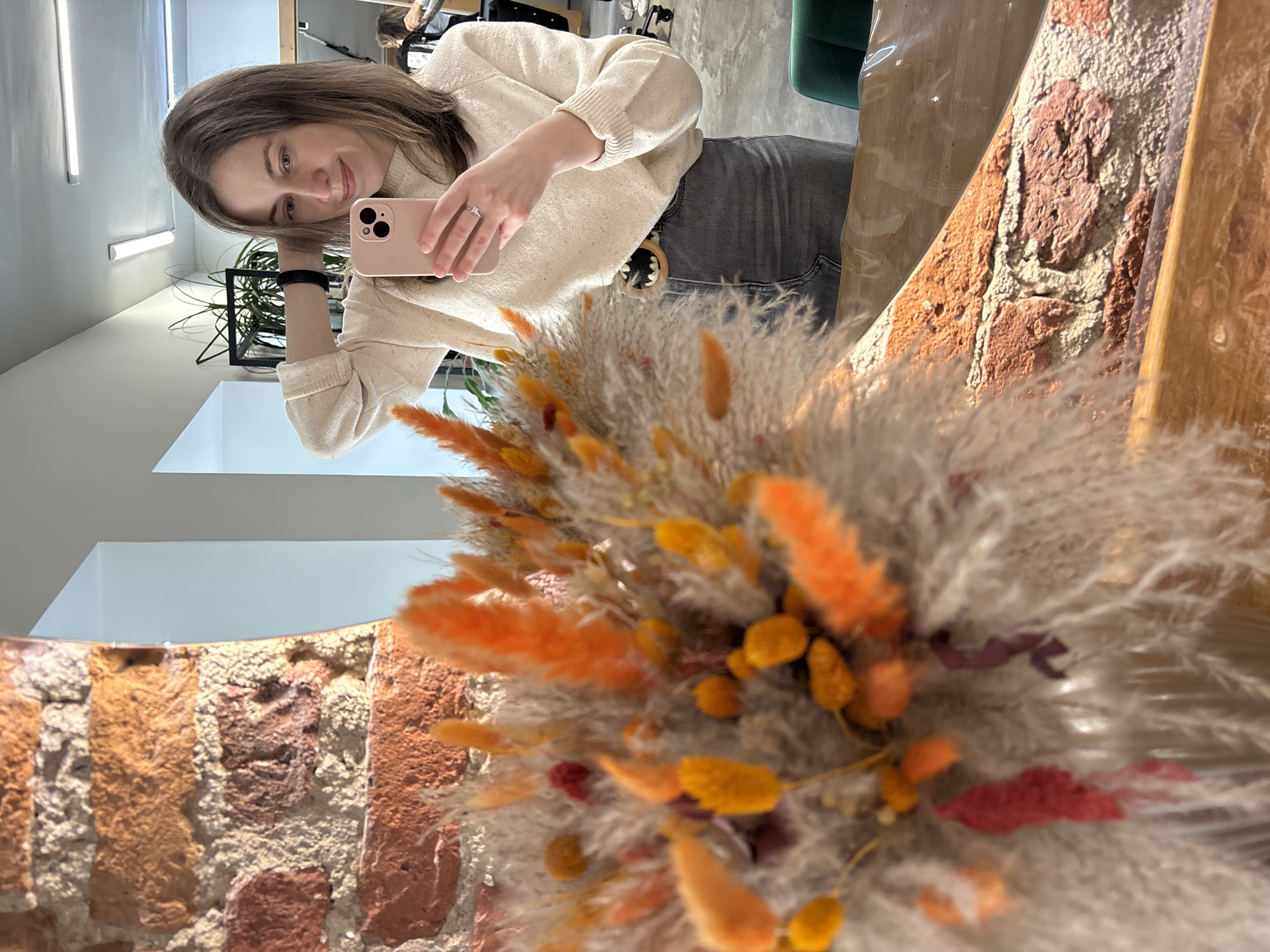 The image shows a woman taking a selfie in a room with exposed brick and modern decor. The woman has brown hair and is wearing a cream-colored sweater and gray jeans. She is holding a pink smartphone. There is a bouquet of dried flowers in the foreground and a mirror behind her, reflecting the room. The location is a modern salon in the city of Saint Petersburg, Russia.