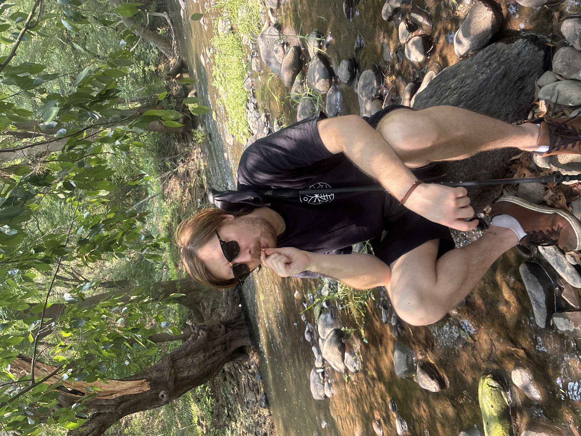A man is sitting beside a stream surrounded by trees and rocks. He is wearing a black t-shirt, black shorts, brown shoes with white socks, and sunglasses. He is holding a thin, dark-colored object (possibly a cigarette or similar item) to his mouth with his right hand. A walking stick is resting on the ground near his legs. The location cannot be confirmed as Cañada de Madero, Mexico without further information.