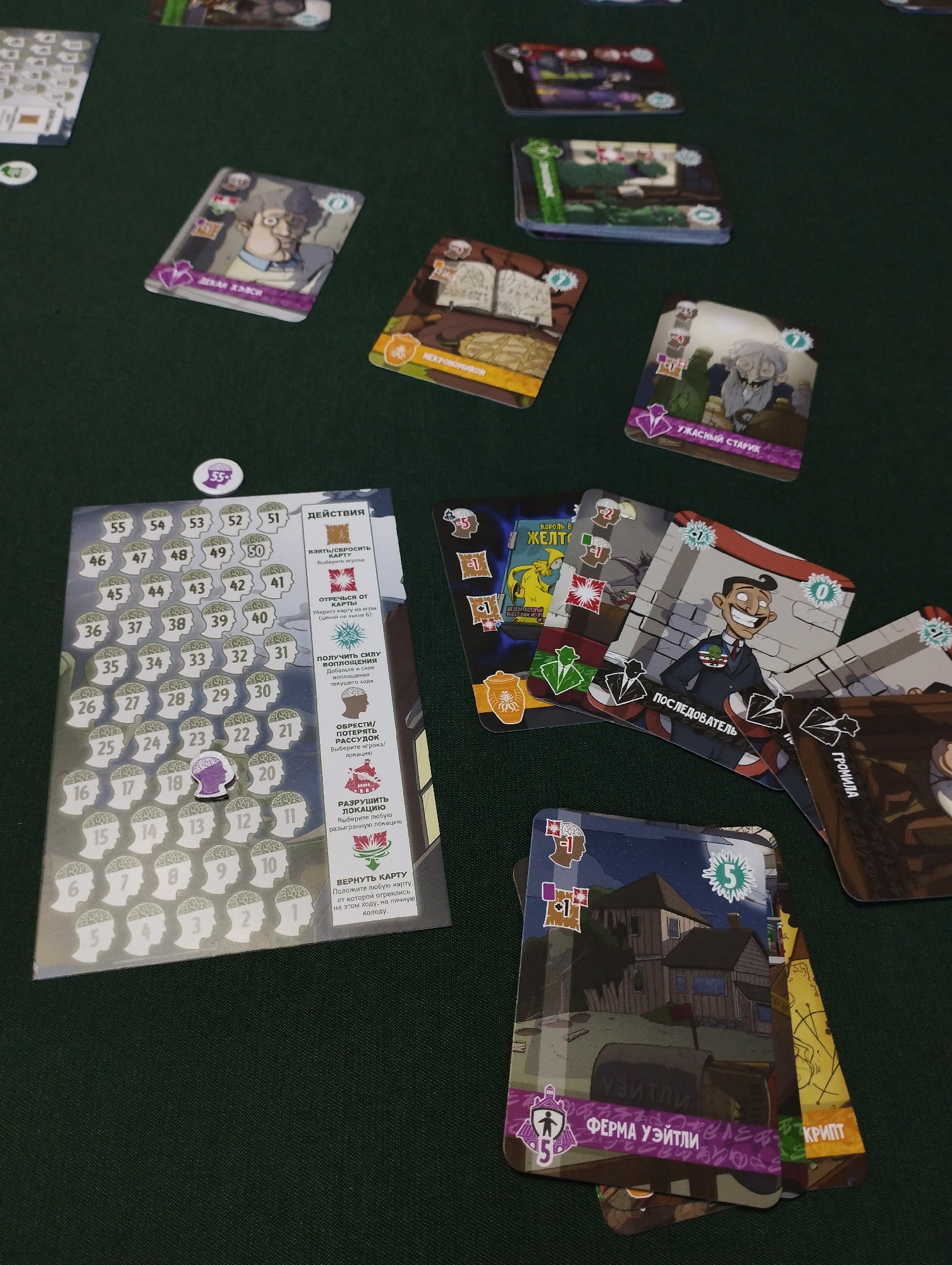 The image shows a tabletop board game laid out on a dark green surface. Several cards are visible, featuring illustrations and Russian text. One card has a grid of numbered circles, with some circles colored in to represent progress. Another card depicts a character with a wide smile wearing a suit and tie, with the text "ПОСЛЕДОВАТЕЛЬ" below. There are also cards with images of houses and abstract designs. A small token with the number 55 is placed on the grid. The environment appears to be indoors, with the focus on the game components. There is no indication of the city of Belaya Kalitva, Russia, or any specific geographical location.