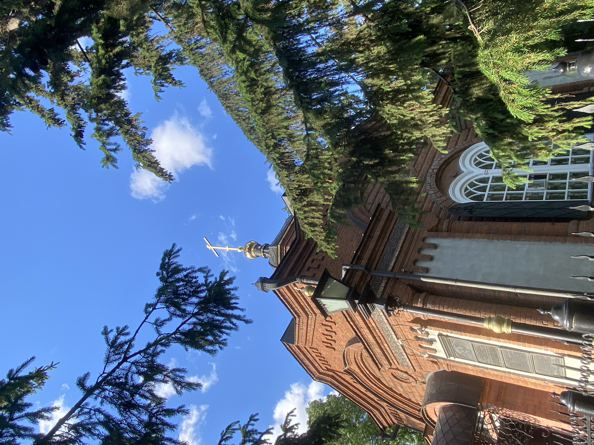 The image shows a low-angle view of the Church on Blood in Yekaterinburg, Russia, framed by trees. The building is brick, with a gold cross atop its dome. A distinct arched window with white frames is visible on the side. The sky is blue with scattered white clouds. A decorative streetlight is attached to the building. The photo appears to have been taken from a street-level perspective.