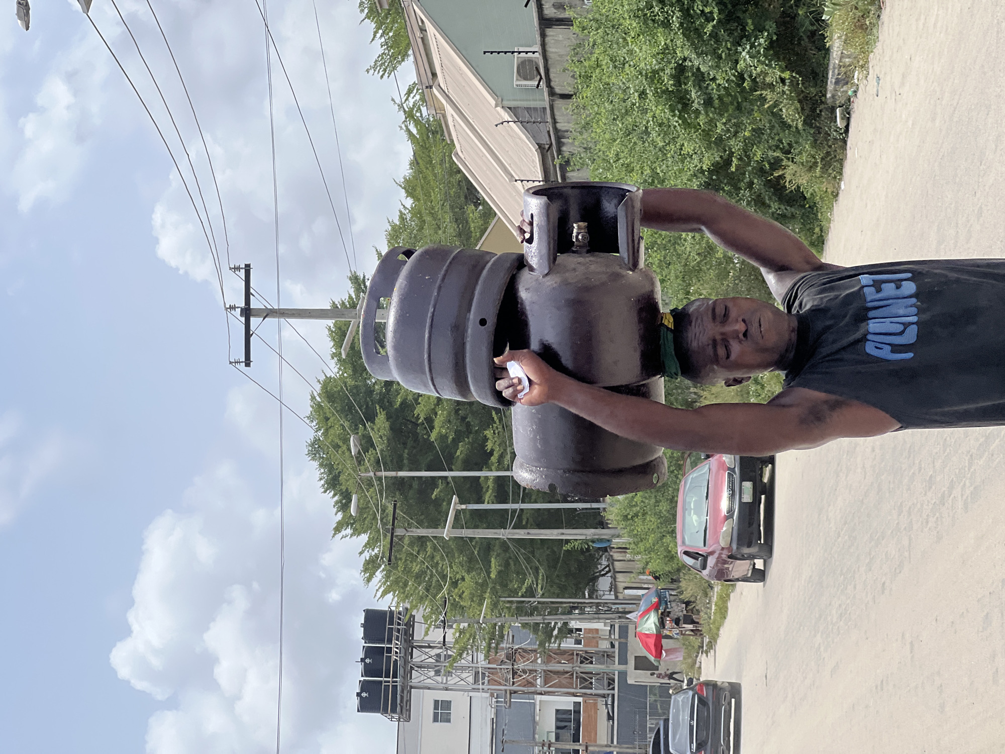 A man is depicted carrying two gas cylinders on a sunny day in Igboefon, Nigeria. The image, oriented vertically, shows the man from the waist up, looking directly at the camera with a neutral expression. He is dark-skinned, has a short haircut, and wears a black t-shirt with the word "PLANET" in blue capital letters across the chest. A green and yellow band is visible around his neck. His arms appear muscular as he holds two dark-colored gas cylinders. One larger cylinder is balanced on a smaller one, which the man grips with both hands.
The scene is an outdoor street environment. In the background, a paved road stretches out with vehicles, including a red sedan on the right and parts of black cars visible. Various buildings line the street, some with light-colored roofs and others surrounded by lush green trees and bushes. One building features large black water tanks mounted on a support structure. Numerous power lines crisscross the sky, supported by wooden and concrete utility poles, some with streetlights attached. The sky is partly cloudy with patches of bright blue, indicating clear weather. The overall impression is one of everyday life and commerce in a semi-urban setting, with the man engaged in the physical labor of transporting gas cylinders.