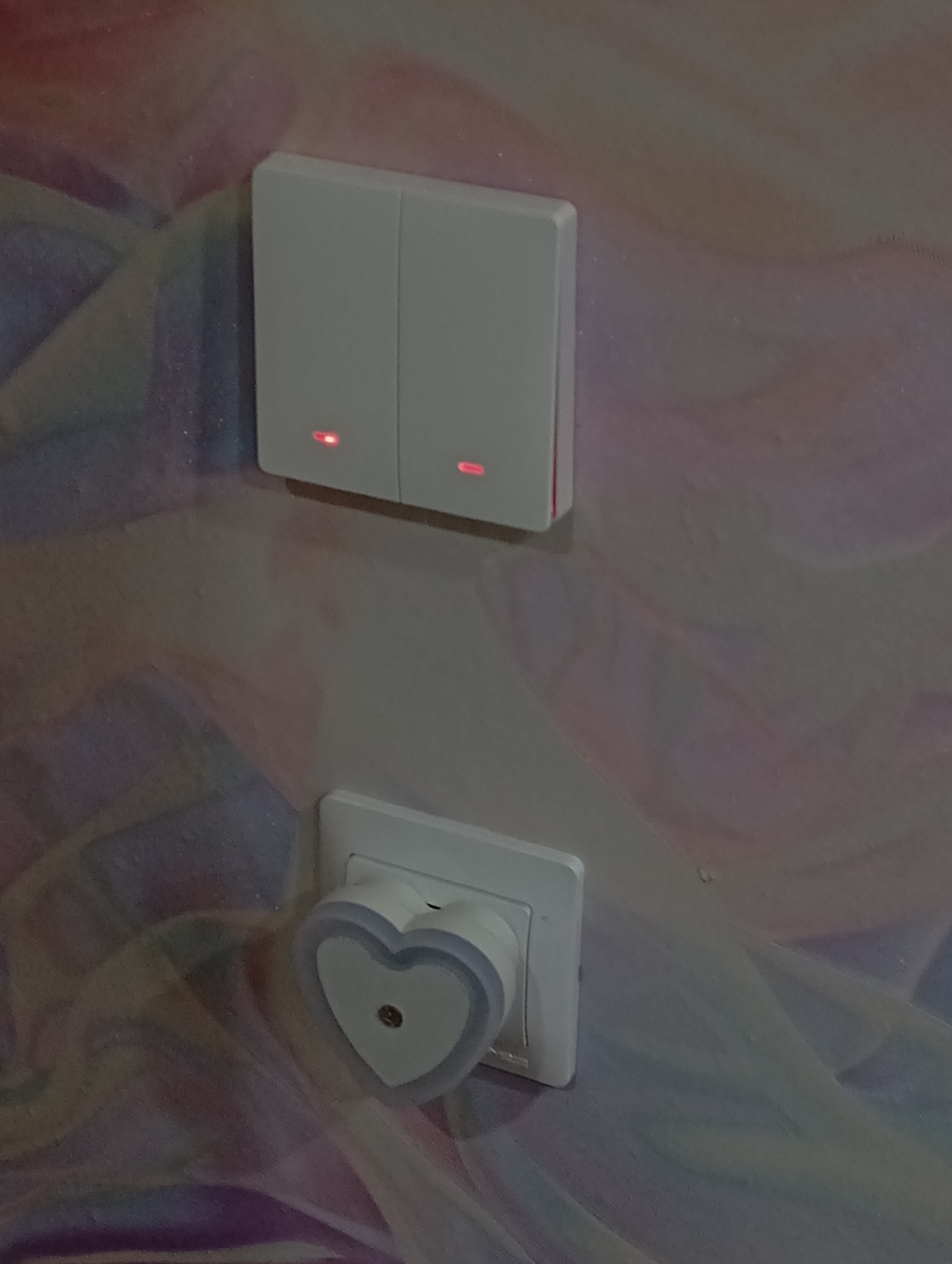 The image shows a close-up of a wall with two light switches and a nightlight. The light switches are white and have red indicator lights. The nightlight is heart-shaped and plugged into an outlet. The wall has a multi-colored design that includes pink, purple, and white. The location and specific details about the city of Krasnodar, Russia, cannot be determined from the visual information provided in the image. There are no people visible in the image, no actions are depicted, and there are no interactions between subjects.