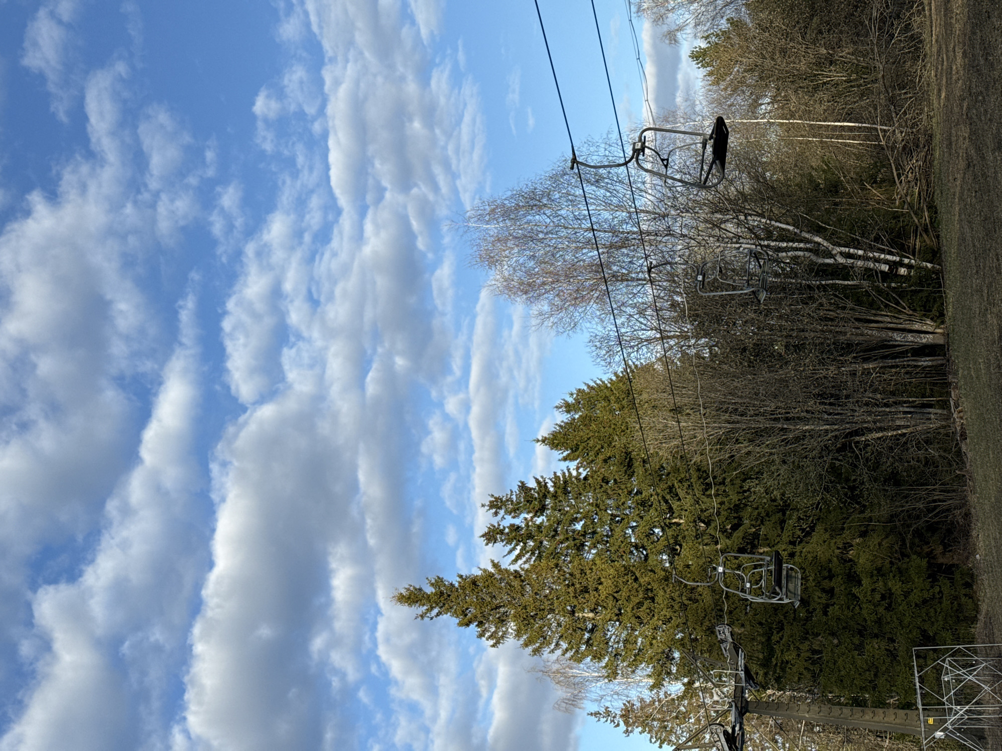 The image shows a ski lift ascending a wooded slope under a blue sky with scattered clouds. The ski lift is the dominant feature, with empty chairs suspended from cables stretching across the frame. Below the lift, a mix of evergreen and deciduous trees covers the hillside. The ground is visible along the right edge of the frame, appearing as a grassy area. The location cannot be definitively determined to be Galevo, Russia based on the image alone.
