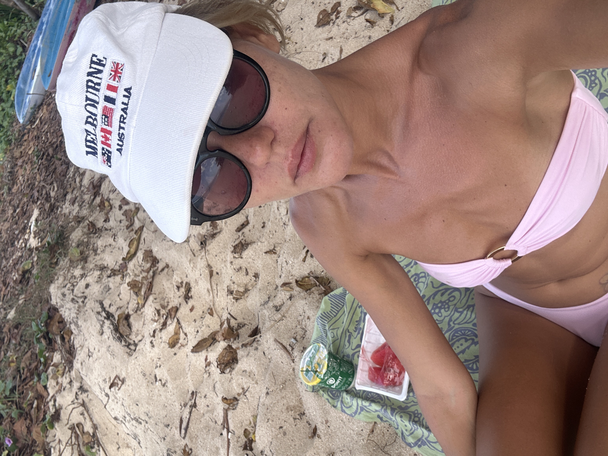 A high-angle, slightly tilted shot shows a person lying on a sandy beach in Kuta Selatan, Indonesia. The person is wearing a white baseball cap with "MELBOURNE AUSTRALIA" embroidered on it, alongside several small flags. They are also wearing sunglasses and a pink bikini. To the right of the person, on a patterned cloth, is a small container of sliced watermelon and a green cup. The background is a sandy beach with some dry leaves and vegetation. The lighting suggests it is daytime. The person appears relaxed.