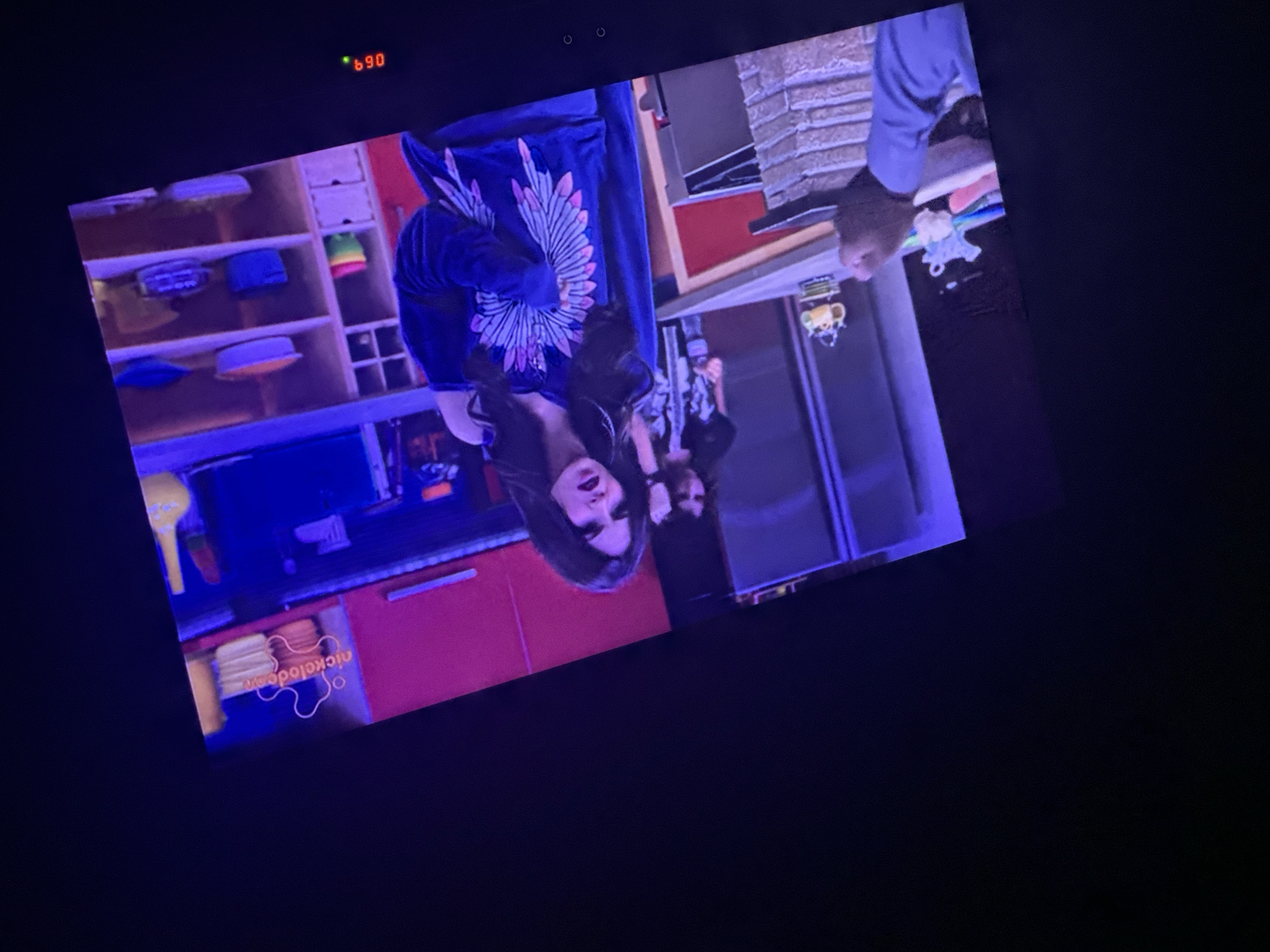 A television screen is showing a scene from a Nickelodeon television show. A female character is in focus, wearing a dark jacket with a floral design on the back. In the background, there is a brightly colored kitchen set with shelving containing various objects. The Nickelodeon logo is visible at the bottom of the screen. A red digital number '690' is in the top left corner. The location is inside a home, likely in Rakovski, Bulgaria.