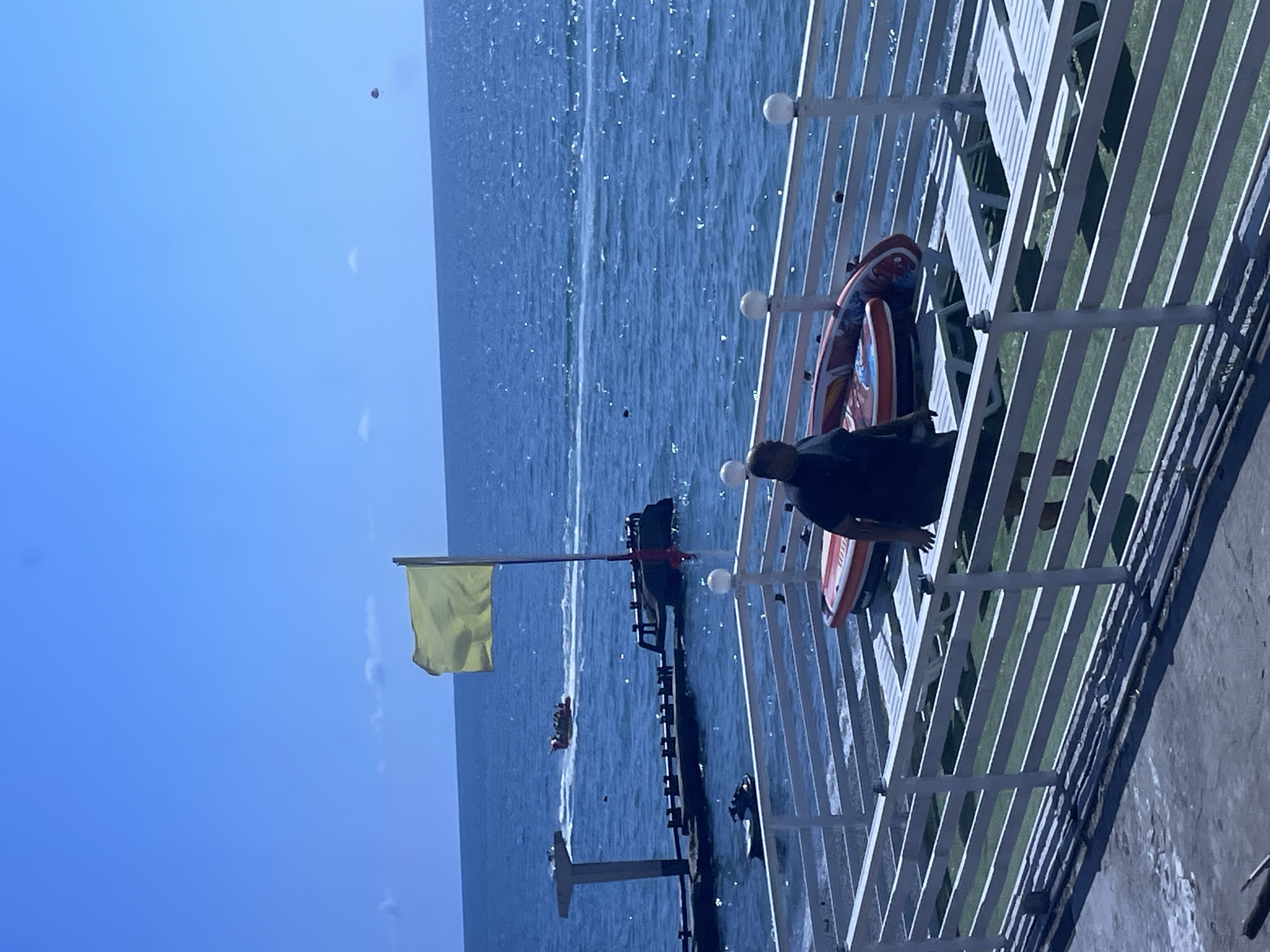 The image captures a sunny day at a beach resort in Sochi, Russia, with the Black Sea stretching out under a clear blue sky. A yellow flag flutters on a pole, possibly indicating good swimming conditions. In the foreground, a person is standing on a white-railed deck overlooking the water, near several lounge chairs and an inflatable raft. The deck appears to be part of a tourist facility.
The middle ground shows the blue expanse of the Black Sea. In the distance, a jet ski is visible near a derelict pier structure. Further out on the water, a banana boat filled with people is being towed by a boat, suggesting recreational activities are taking place. A person can be seen paragliding high in the sky above the sea, another indication of leisure pursuits. The overall scene conveys a lively atmosphere at a seaside location in Sochi.