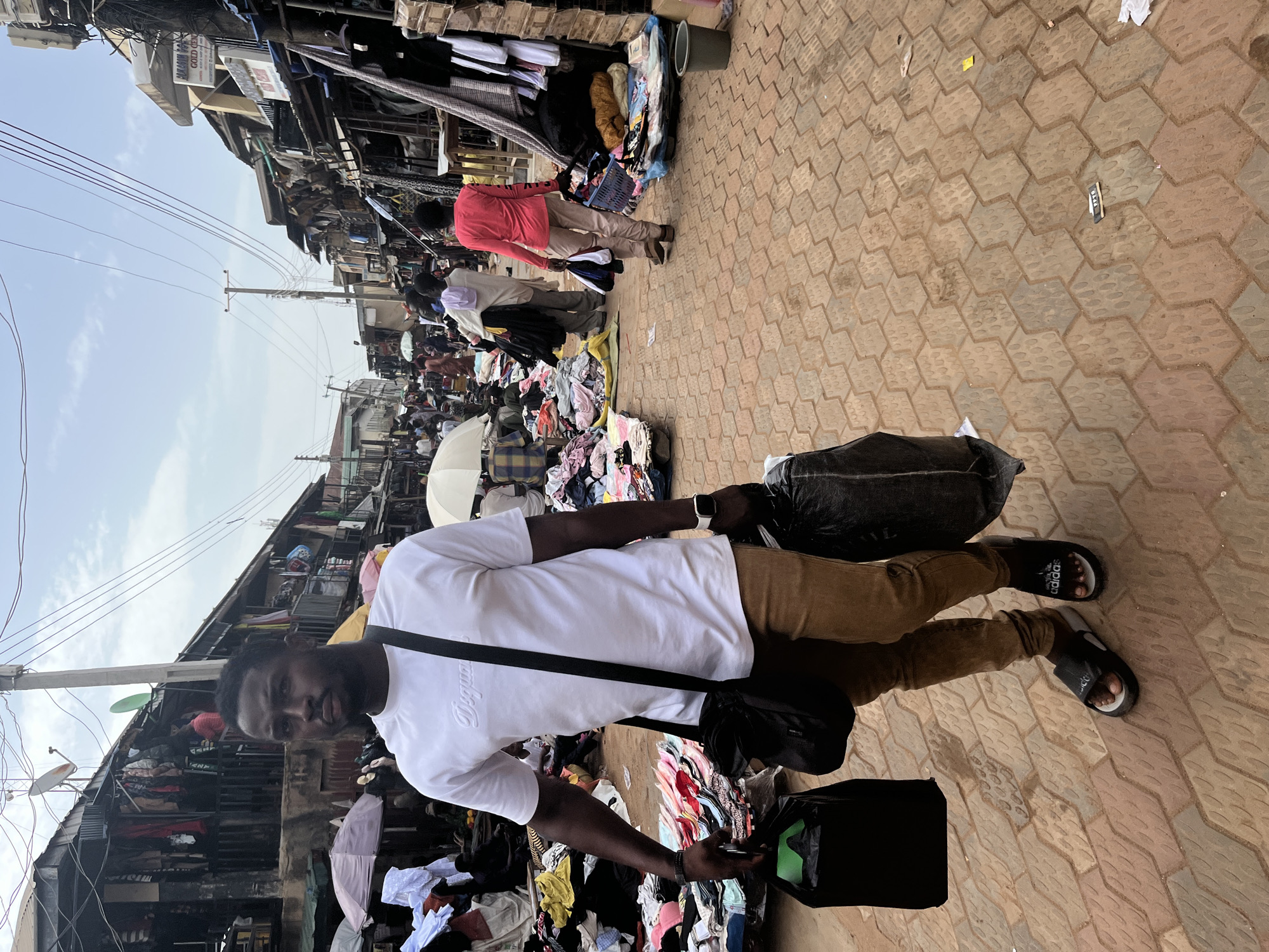 This is a street-level, medium-shot photograph taken in the city of Jos, Nigeria, depicting a bustling market scene. The foreground is dominated by a young Black man walking towards the viewer, carrying two large black plastic bags, one in each hand. He is wearing a white t-shirt with a subtle embossed logo, brown trousers, and black slide sandals. A black messenger bag is slung across his chest. His gaze is directed forward, and he appears to be walking through a crowded marketplace.
The background reveals a vibrant and somewhat chaotic market environment. Stalls and makeshift shops line both sides of the narrow street, displaying a wide array of goods, primarily clothing, which are spread out on tarps or hanging. Umbrellas are visible, providing shade for vendors or shoppers. Several other people are seen scattered throughout the market, some browsing, others engaged in transactions. The ground is paved with patterned interlocking bricks. Overhead, a complex network of electrical wires crisscrosses the sky, a common sight in urban Nigerian settings. The architecture of the surrounding buildings appears somewhat weathered and densely packed. The overall impression is one of an active and lively commercial hub within Jos.