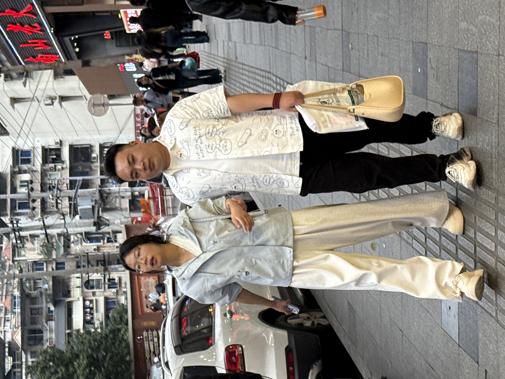 Observed in the Liangjiang district of China, a man and a woman stand together on a paved street. The man wears a white shirt patterned with cartoon drawings, black pants, and white sneakers, while holding a tan bag and a white bag with chopsticks. The woman, wearing glasses, is dressed in a light blue jacket and tan pants, with matching sneakers. They stand in front of a white car parked near buildings and vegetation, with other pedestrians and shops visible in the background.