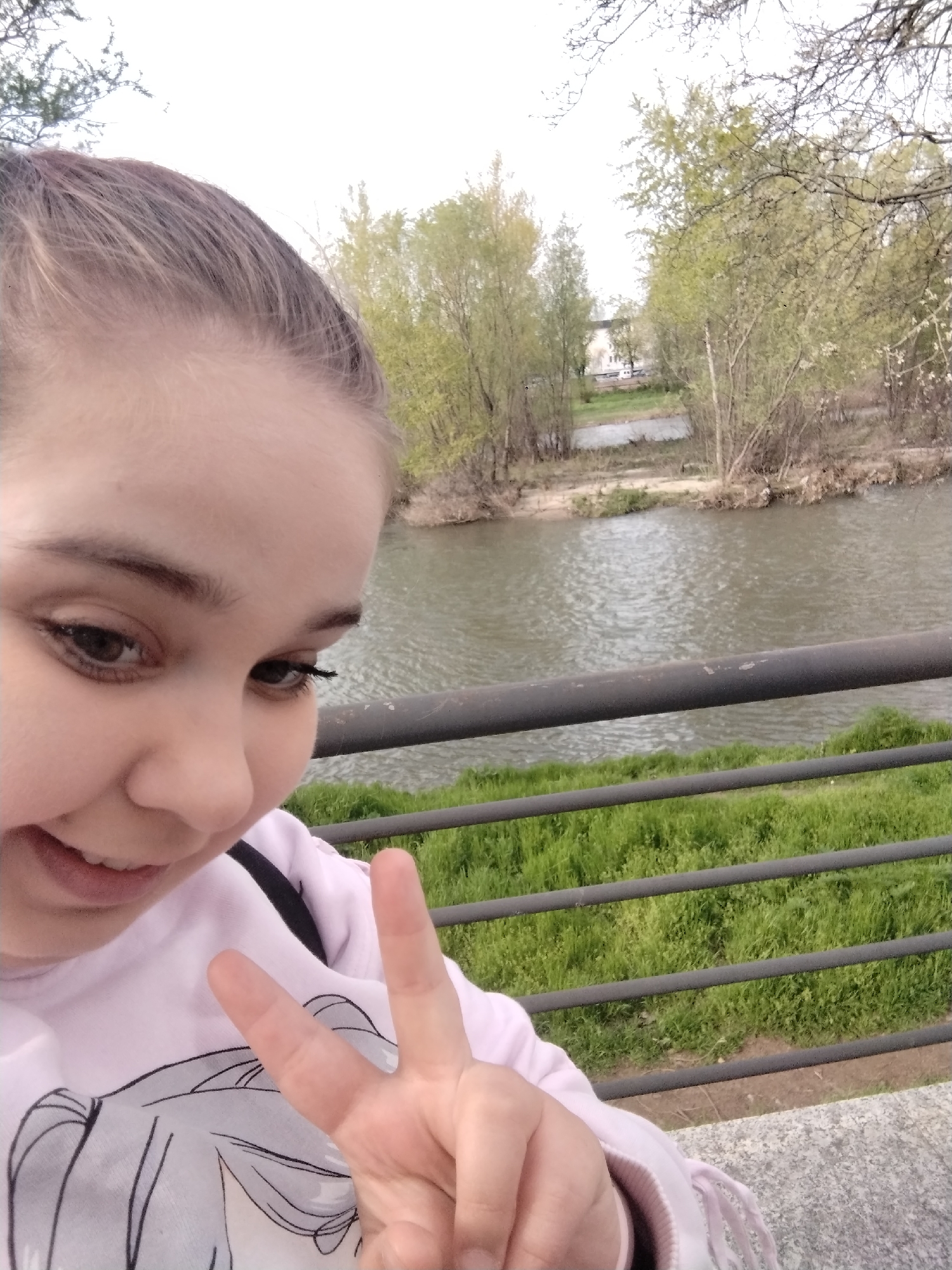 A person is taking a selfie in Plovdiv, Bulgaria. The person is a fair-skinned female with brown hair pulled back from her face. She is wearing a light pink sweatshirt. She is holding up her hand in a peace sign. Behind her is a river with trees on the opposite bank. A metal railing is visible in the foreground. Green grass grows below the railing. A building can be seen through the trees on the opposite bank.