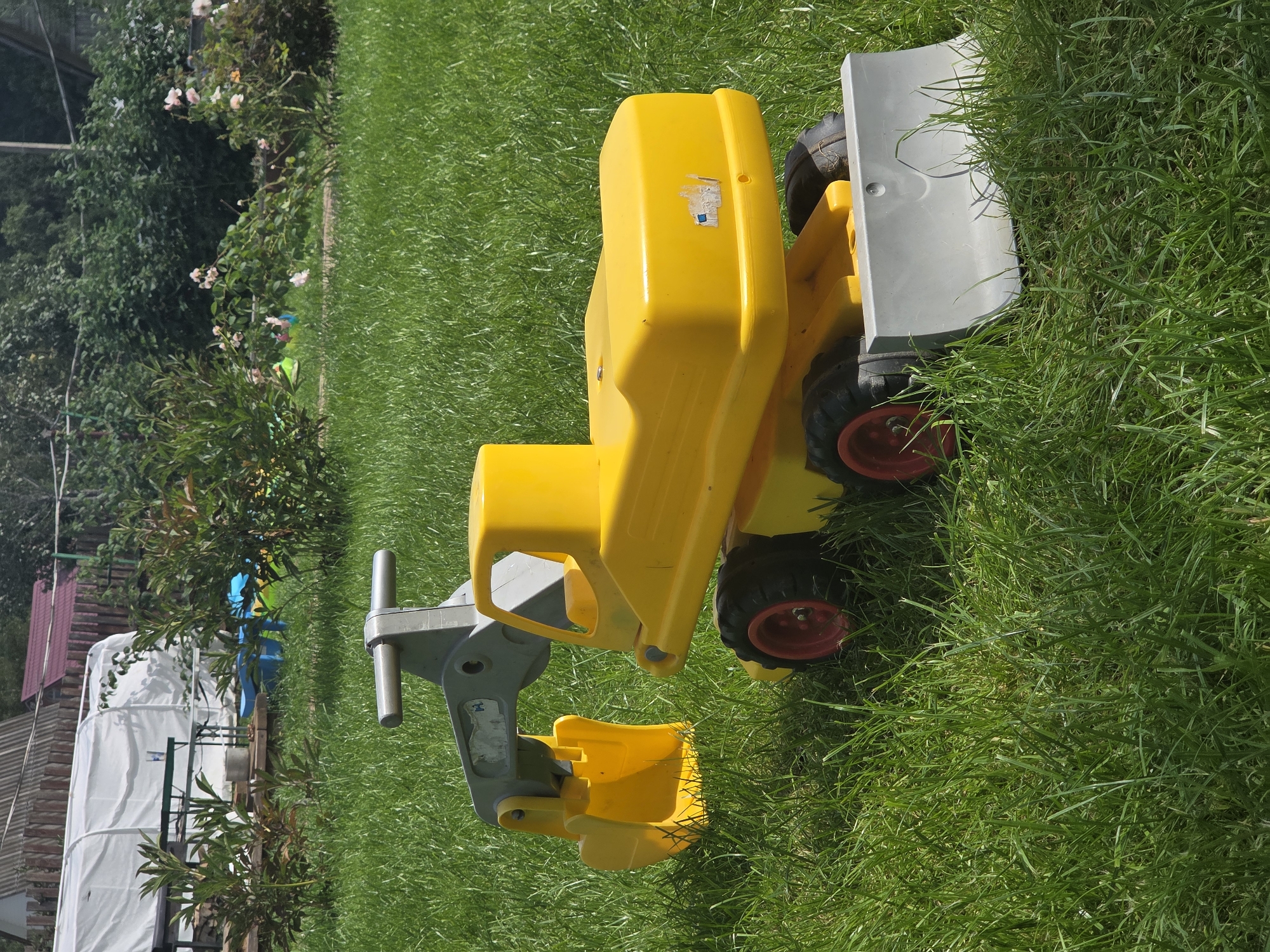 A yellow toy excavator with a gray arm and bucket sits on lush green grass in the backyard of a house in Volgograd Oblast, Frolovo, SNT Voshod, Russia. The toy is positioned to the left of the frame, with its front facing right. In the background, a white greenhouse and a wooden fence are visible, along with some rose bushes and other vegetation. The lighting suggests it is a sunny day.