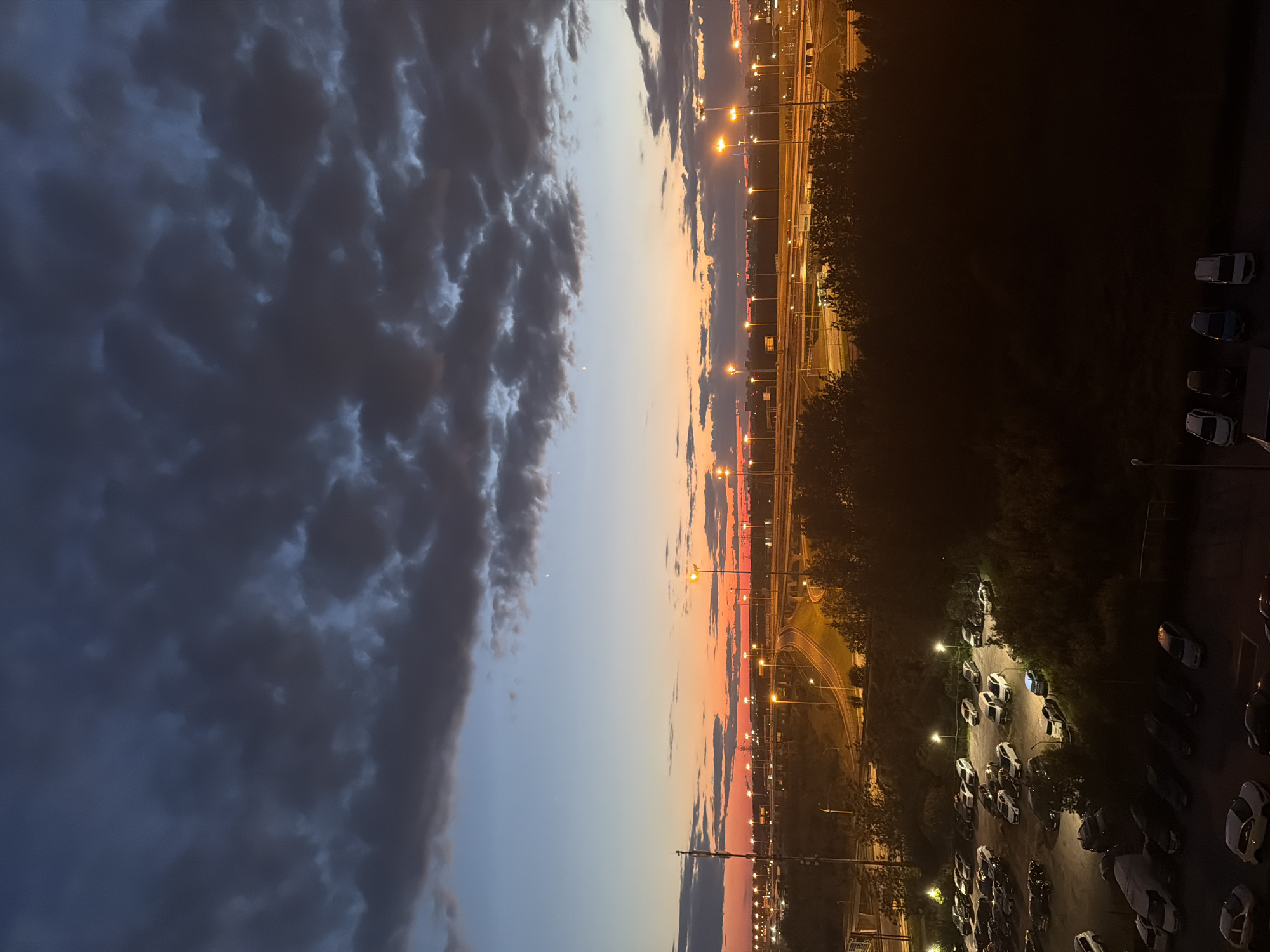 The image depicts a twilight scene over a suburban or industrial area. The sky is a gradient of deep blue to orange as the sun sets, with dramatic dark clouds overhead. Below the sky, a highway interchange with multiple lanes and on-ramps is illuminated by streetlights. Trees and vegetation line the roads and the foreground. In the middle ground and foreground, there are several parking lots filled with cars. The scene is largely devoid of human presence.
