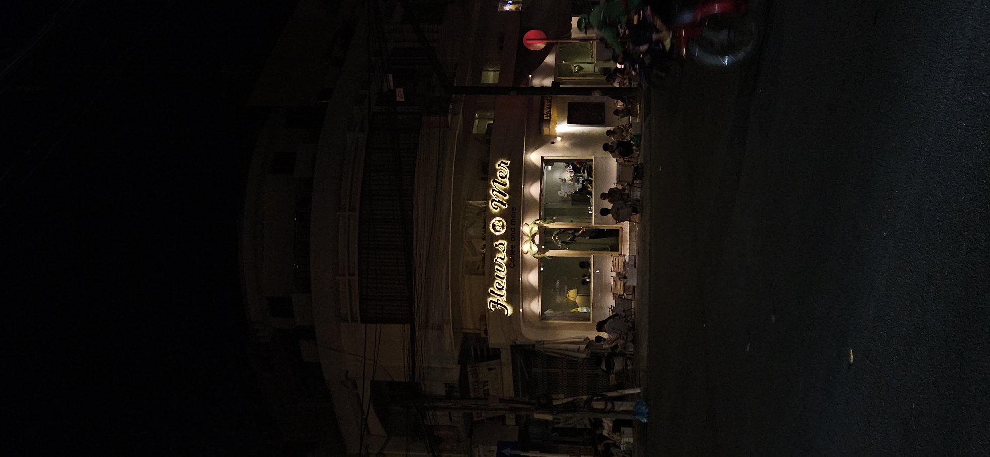 The image captures a vibrant night scene along a street in Nha Trang, Vietnam. The primary subject is a brightly lit storefront, likely a café or restaurant, standing out against the dark urban environment. The establishment's name, "Fleurs @ Mer," is prominently displayed in elegant, golden, illuminated script, with the tagline "Coffee and more" visible below it.
Inside the well-lit interior, visible through large glass windows, several patrons can be seen. An outdoor seating area is set up on the sidewalk directly in front of the storefront, where people are seated at tables and chairs, enjoying the evening. On the street to the right, a motorbike is partially visible, with a person wearing a green helmet and jacket, suggesting a delivery rider or commuter. A red circular traffic sign can be faintly seen in the upper right quadrant. The rest of the scene is enveloped in darkness, characteristic of nighttime, with only faint outlines of other buildings or overhead wires visible on the left. The image portrays a lively yet relaxed urban evening in Vietnam.