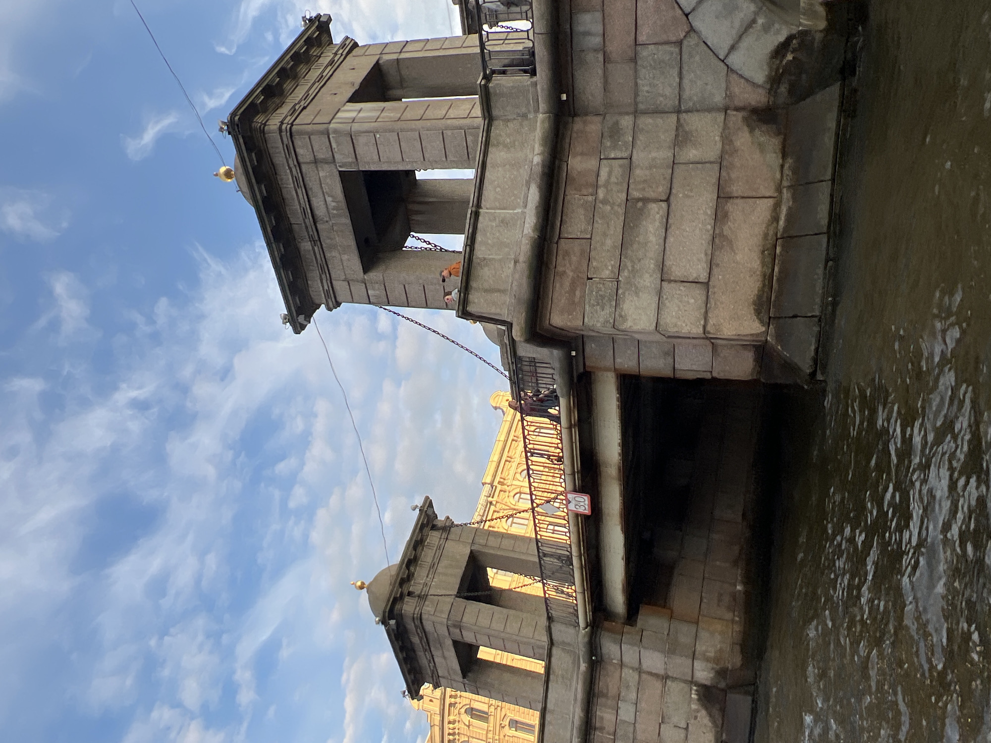 This is a medium shot, captured from a low angle, of a stone bridge in Saint Petersburg, Russia. The bridge features two symmetrical stone towers with domed tops, each topped with a golden sphere. Heavy chains connect the towers to the bridge's deck. The stone masonry of the bridge is rough-hewn and appears weathered.
On the bridge's deck, a person in dark clothing walks away from the viewer towards the right side of the frame. Further back, two individuals are visible on the bridge's walkway. The bridge spans a dark, murky body of water, likely a canal or river, with ripples on the surface.
In the background, to the left, a large, ornate building with a yellow facade and arched windows is visible, characteristic of St. Petersburg's architecture. The sky above is a bright blue, dotted with wispy white clouds. A speed limit sign indicating "30" is attached to the railing of the bridge. The overall lighting suggests it is late afternoon or early evening, with the sun casting a warm glow on the buildings.