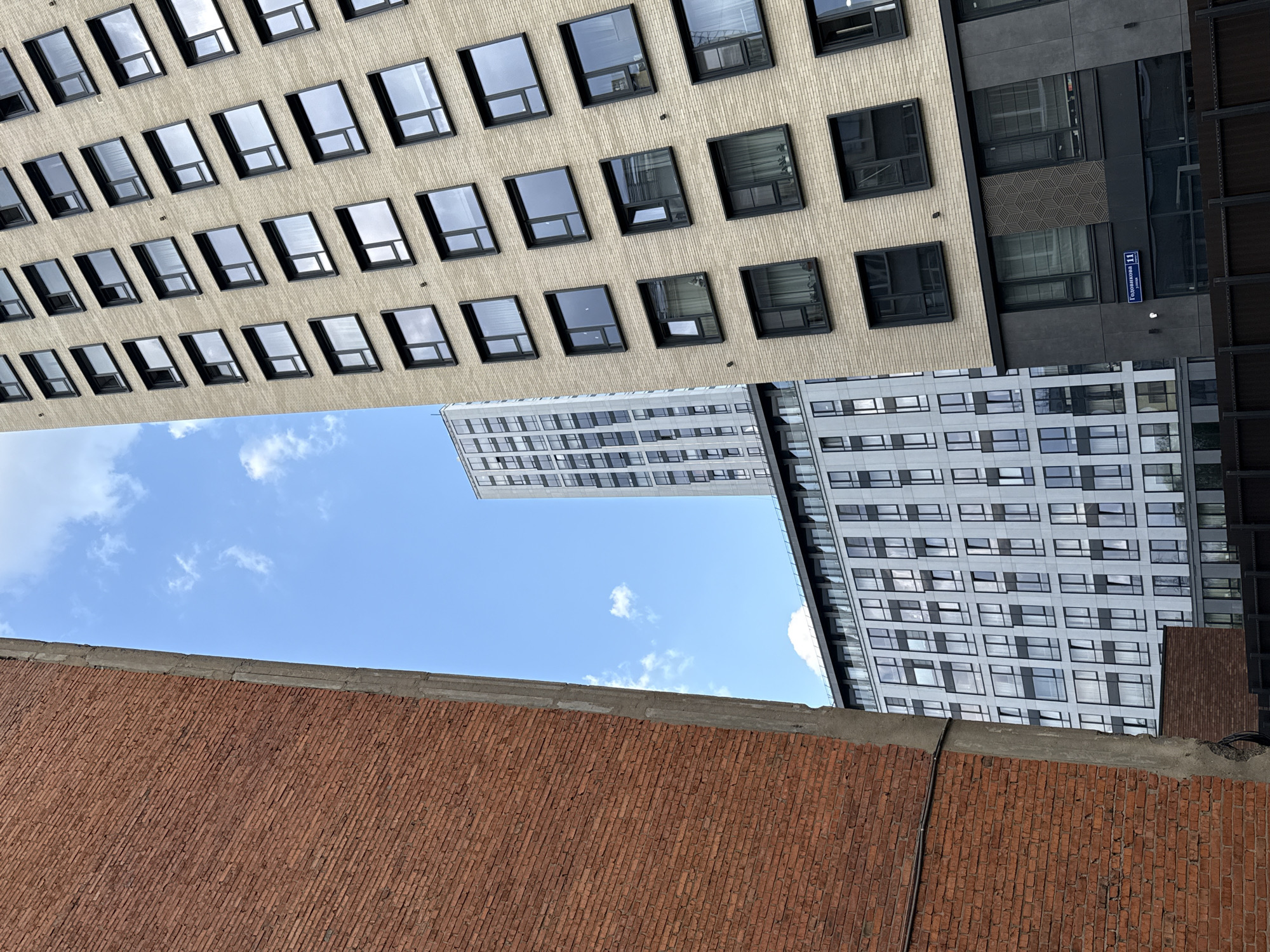 The image shows a view of the sky framed by buildings in Khimki, Russia. At the bottom is a brick wall, likely part of a lower building. Above it, the sky is visible with some clouds. Surrounding the sky are parts of three buildings with various architectural styles: one with light-colored brick and black-framed windows, one with grey panels, and another with a mix of grey panels and windows. A sign reading "Героев Панфиловцев 11" (Heroes of Panfilov 11) can be seen on one of the buildings. The perspective suggests the photo was taken from a lower vantage point, looking upwards.
