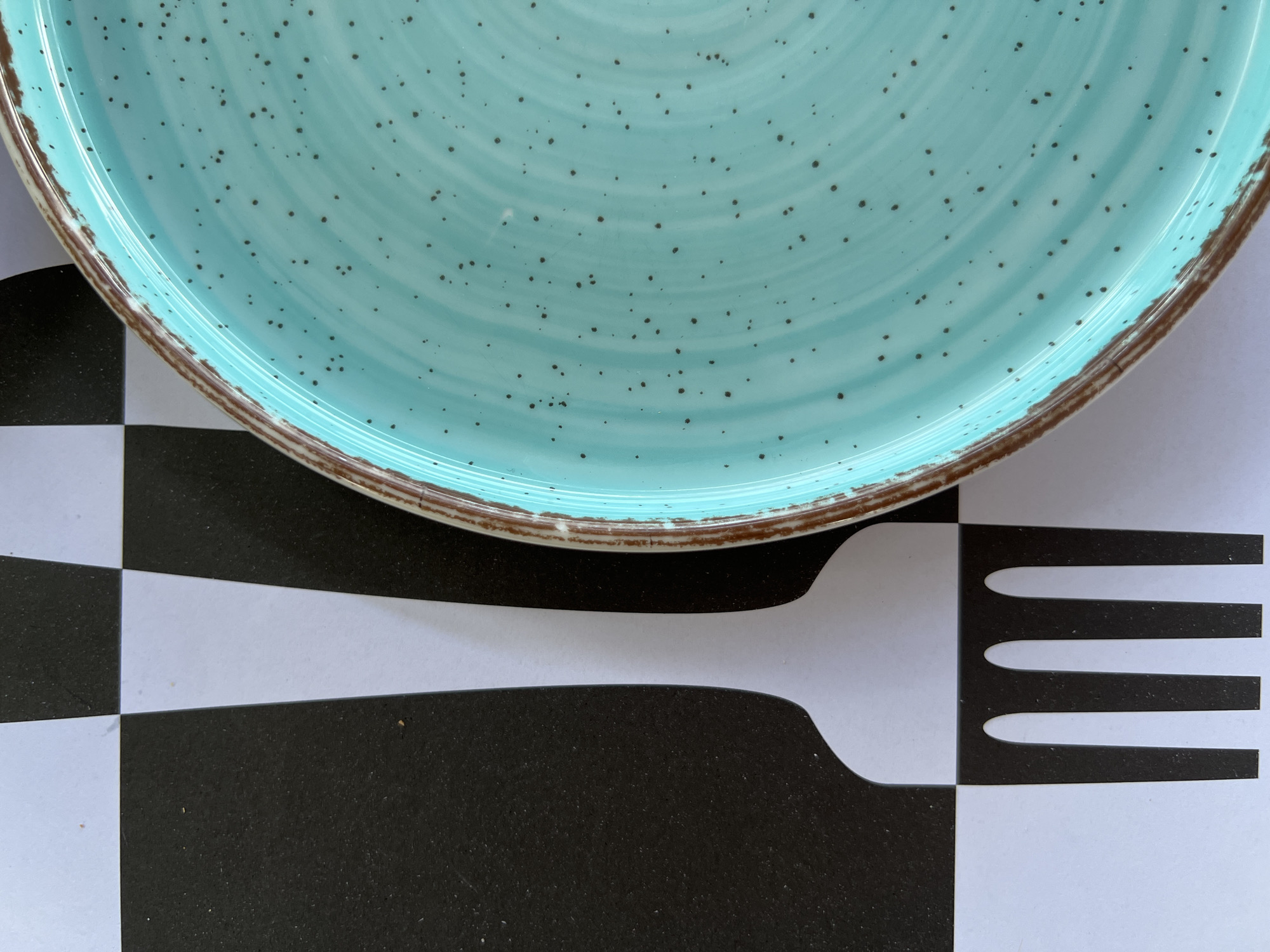 An overhead shot of a turquoise plate with brown trim, speckled with dark spots, rests on a black and white checkered surface. The checkered surface resembles a placemat with a large, stylized fork imprint in white and black. The plate is positioned in the upper right quadrant of the image, partially overlapping the checkered pattern. The fork imprint is centrally located and oriented vertically. The scene appears to be indoors, possibly in a restaurant or home setting, located in Eretria Municipal Unit, Greece. There are no people or active interactions depicted.