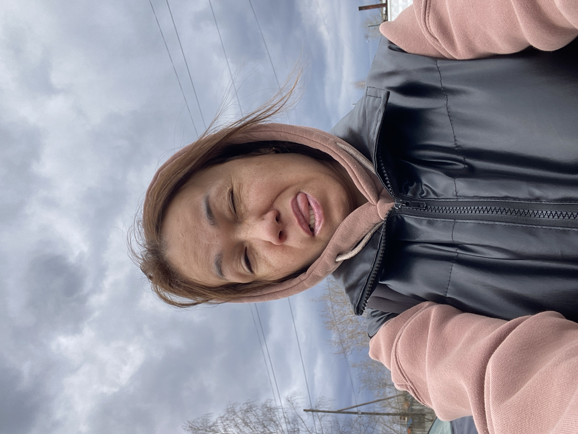 A woman is centered in the frame, sticking her tongue out with her eyes closed. She's wearing a pink hoodie underneath a black puffy vest. The sky is overcast with powerlines visible in the background. Bare tree branches suggest it is either late autumn or early spring. The location is likely in the city of Chaikovsky, Russia.