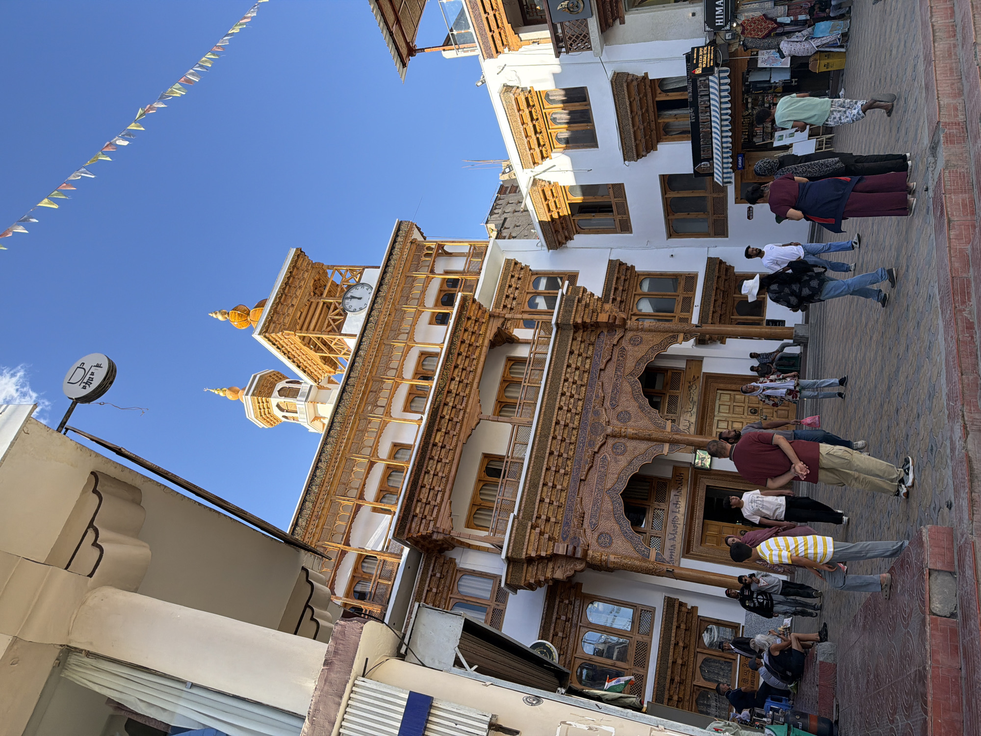 The image shows a street scene in Leh, Ladakh, India, featuring the Jama Masjid. The architecture is distinctive, with white walls and ornate wooden carvings and latticework adorning the balconies and entrance. Several people are visible in the foreground and middle ground, some walking, some standing, and one person sitting. The sky is clear and blue. In the background, more buildings with similar architectural styles are present, along with shops. Flags are strung across a street in the upper right portion of the image. The scene appears to be a bustling public area.