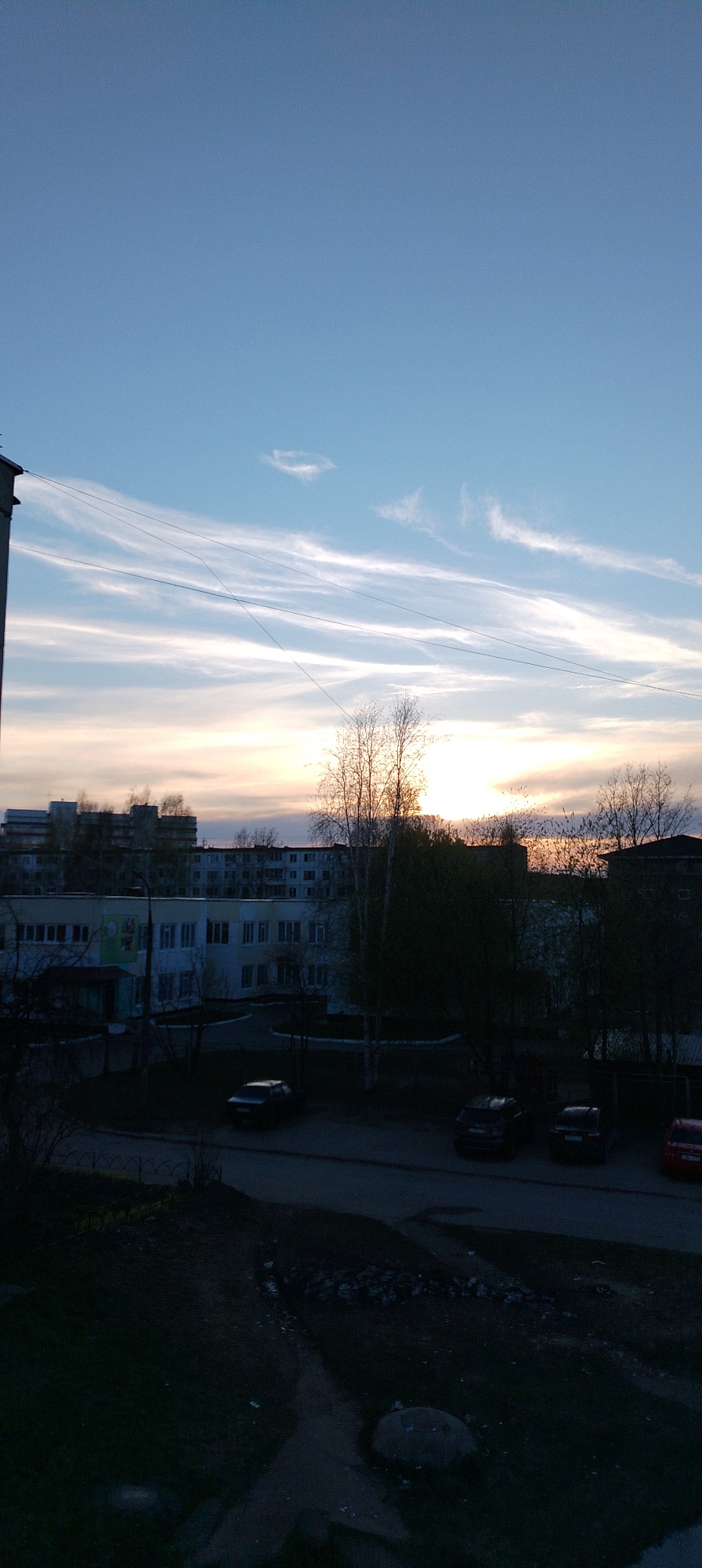 The image captures a cityscape at dusk. The sky is a gradient of blue and orange, indicating a sunset. Wispy clouds streak across the sky. Buildings are visible in the midground, including a white building with what appears to be a mural or sign on the side. Trees obscure some of the buildings. A row of parked cars lines the street. The foreground is mostly dark and undefined. The location is Unknown, Unknown.