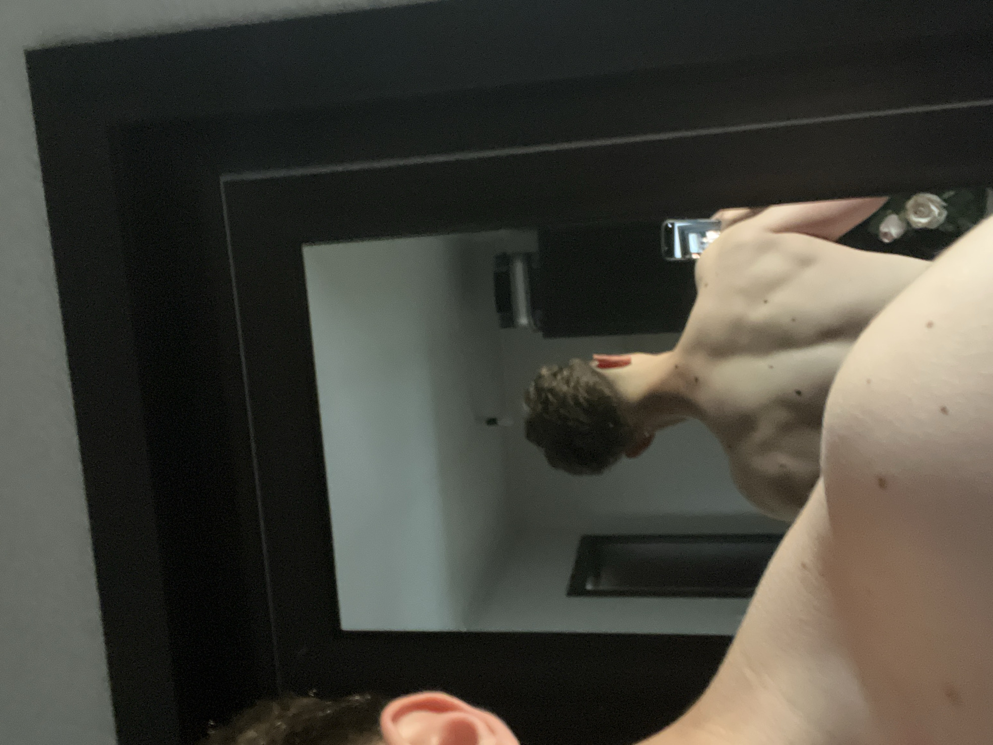 The image is a reflection in a mirror with a thick, dark frame. In the reflection, a person is visible, partially nude, holding a mobile phone in their right hand, presumably to take the picture. The person has short brown hair, and their back is turned toward the camera. The reflection also shows a room with a wall and a rectangular object on the wall. The location in the city of Zlatograd, Bulgaria cannot be determined from this image.