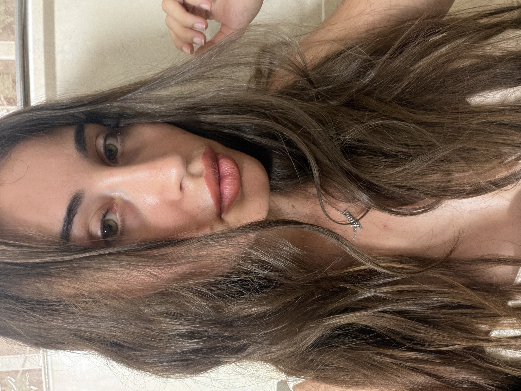 A close-up, head-and-shoulders portrait of a young woman with long, wavy brown hair is captured in what appears to be a bathroom in Tersefanou, Cyprus. She is looking directly at the camera, her head slightly tilted. Her makeup includes lipstick and possibly eyeshadow. She is wearing a necklace with a pendant spelling "Mom". Her right hand is visible, touching her hair. The background consists of tiled walls.