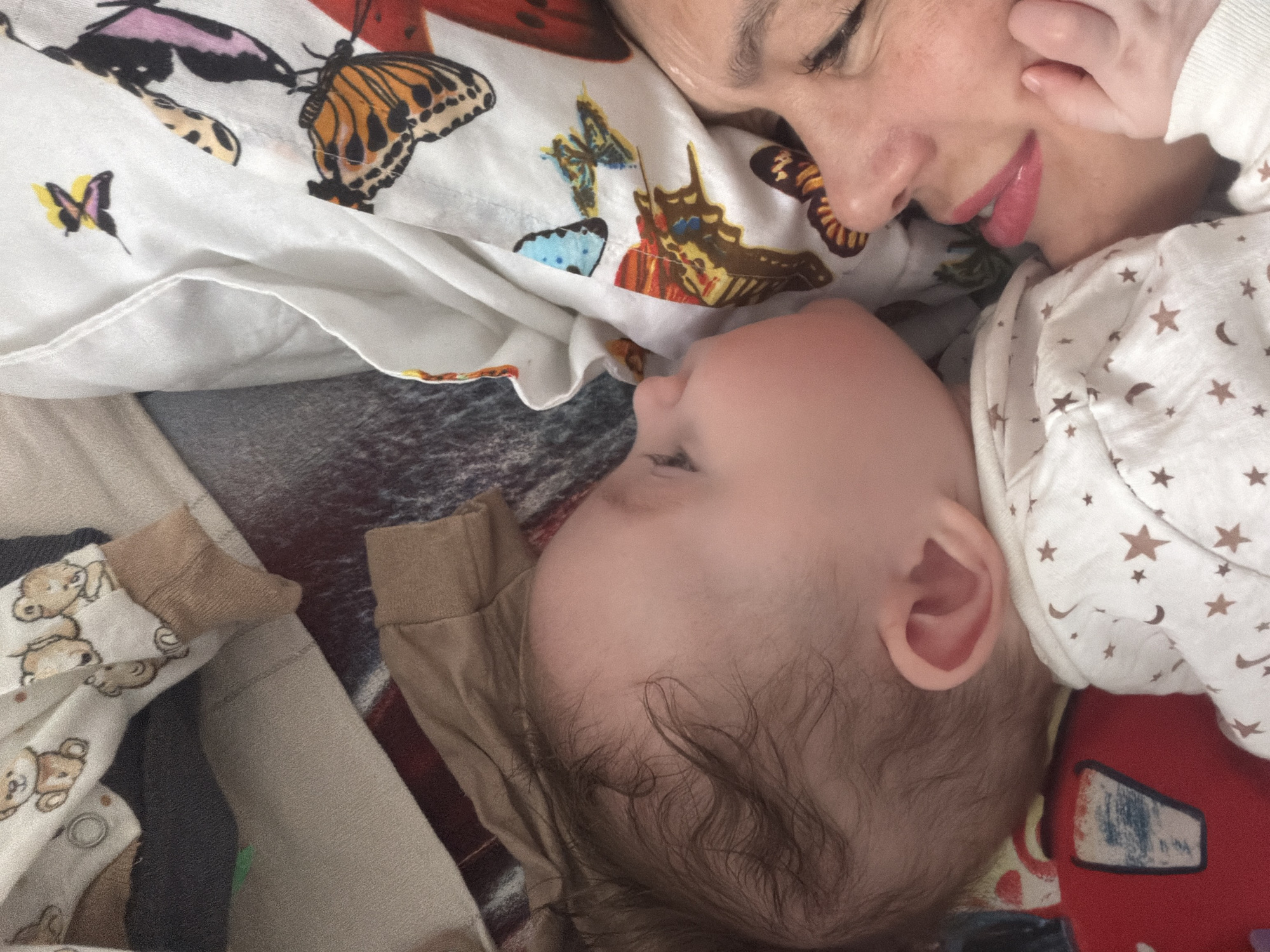A close-up photograph shows a woman and a baby lying down. The woman, facing upwards and to the right, has her eyes closed and is partially obscured by a white garment patterned with butterfly designs. The baby, positioned below the woman and facing left, is also lying down. The baby's head is visible, with wisps of dark hair. The baby is partially covered by a white garment with brown stars and moons, and a red object with a white design. To the left of the baby is a pile of folded baby clothes, some patterned with teddy bears. The setting appears to be indoors. There is no visible information to confirm it is Chaikovsky, Russia.