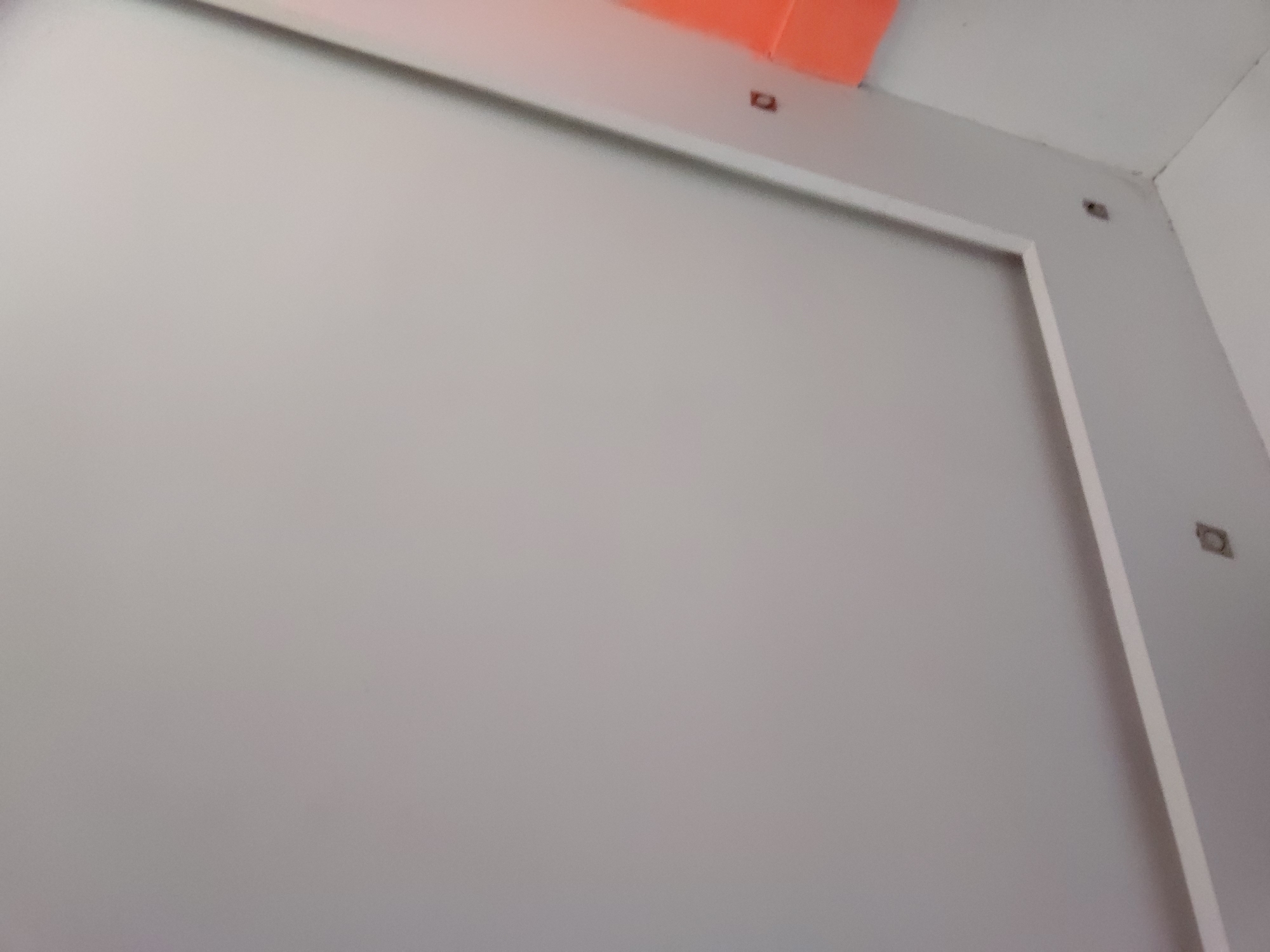 An interior shot focuses on a plain, light grey wall or large panel. This dominant grey surface is framed along its top and right edges by a lighter grey or white trim. In the upper-left portion of the image, an orange object or surface is partially visible, contrasting with the neutral tones of the room. Small, dark, square-shaped elements are embedded within the trim and the adjacent wall; two are seen along the top frame and two along the right side, suggesting fasteners or possibly electrical outlets. The top-right corner of the image reveals a clean intersection of two white ceiling or wall surfaces. The lighting is diffused and even, indicating an indoor setting without direct sunlight. There are no people, discernible activities, or visible text. The scene depicts a static architectural detail, characteristic of an unfinished or minimalist interior. The location context is Santa Ana, Venezuela.