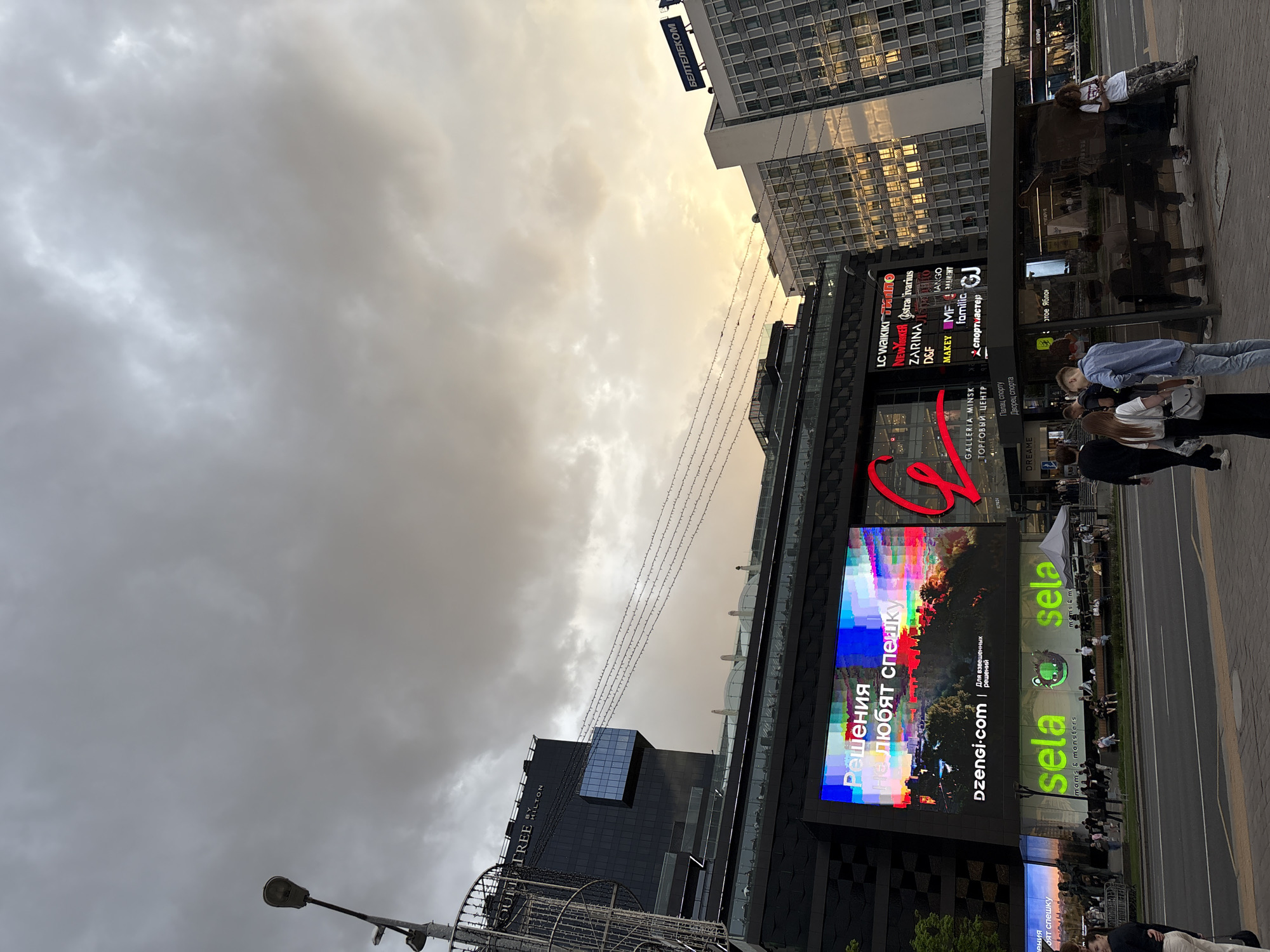 The image shows a street scene in Minsk, Belarus, with modern buildings and digital billboards dominating the view. A tall building with a "BELTELECOM" sign is visible. The billboards advertise brands like "Sela" and "Dzengi.com." People are walking on the sidewalk. The sky is overcast with dramatic cloud formations. A bus stop with waiting people is present on the right side of the street. Additional businesses such as "LC Waikiki" and "Zarina" are advertised on the building fascia. A lower structure displays brands available inside the mall. The "DoubleTree by Hilton" hotel is also visible. Street lighting is present.