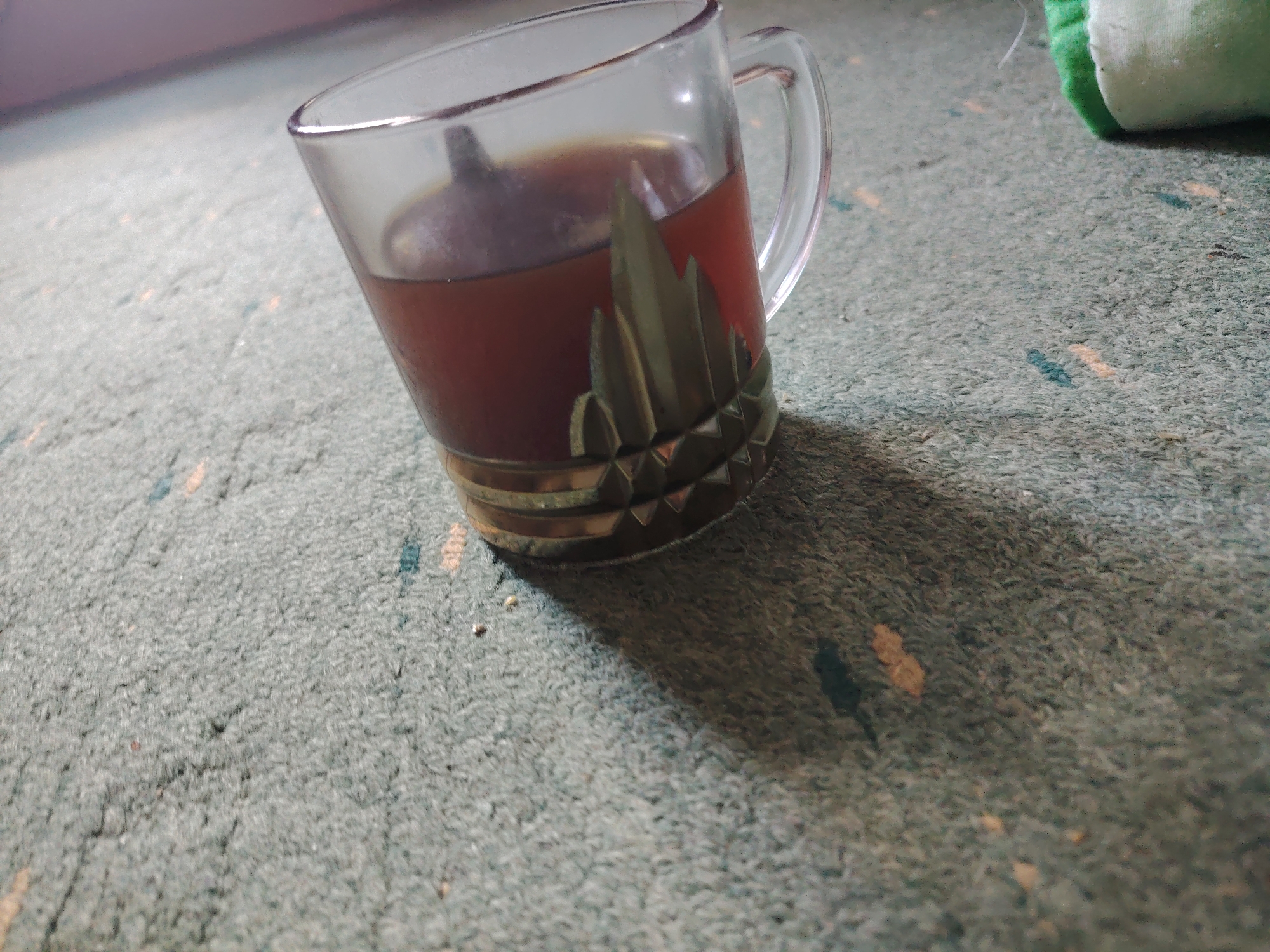 A clear glass mug filled with dark liquid, likely tea or coffee, rests on a textured, greenish-gray carpet. The mug has a metallic decorative band around its base, featuring a repeating geometric pattern that resembles stylized mountain peaks. The mug is tilted slightly to the right, casting a shadow on the carpet. In the upper right corner of the image, a blurry green and white object is partially visible. The carpet has faint, scattered flecks of color, appearing as small yellow and blue dots. The background is out of focus, suggesting a domestic indoor setting. There is no indication of the city of Moscow, Russia, or any specific location. The scene depicts a solitary mug on a carpeted surface.