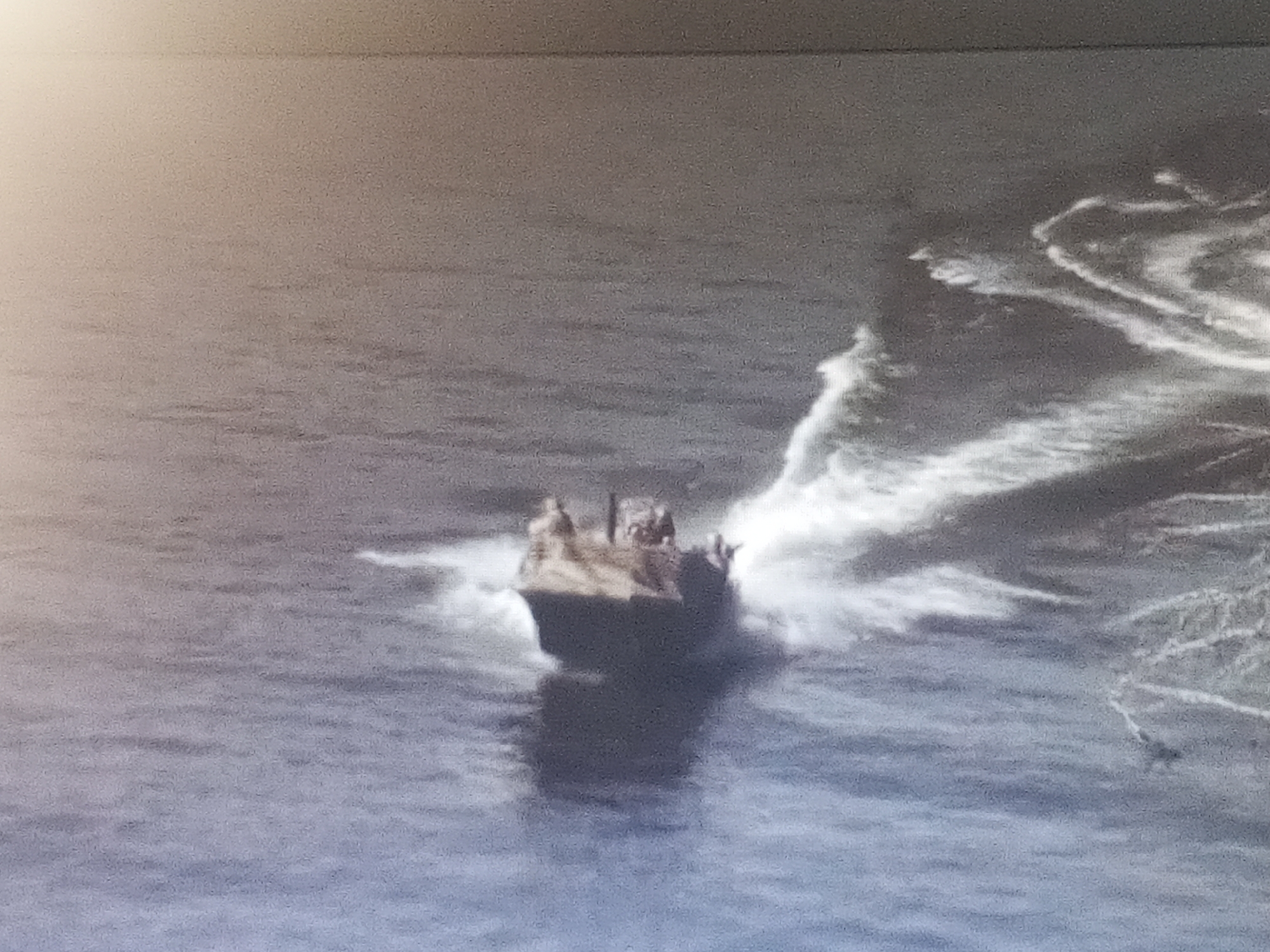 The image shows a military landing craft approaching a shore. The vessel is moving through the water, creating a large wake. There are personnel visible on the landing craft. The location is unspecified. The environment appears to be an ocean or sea.