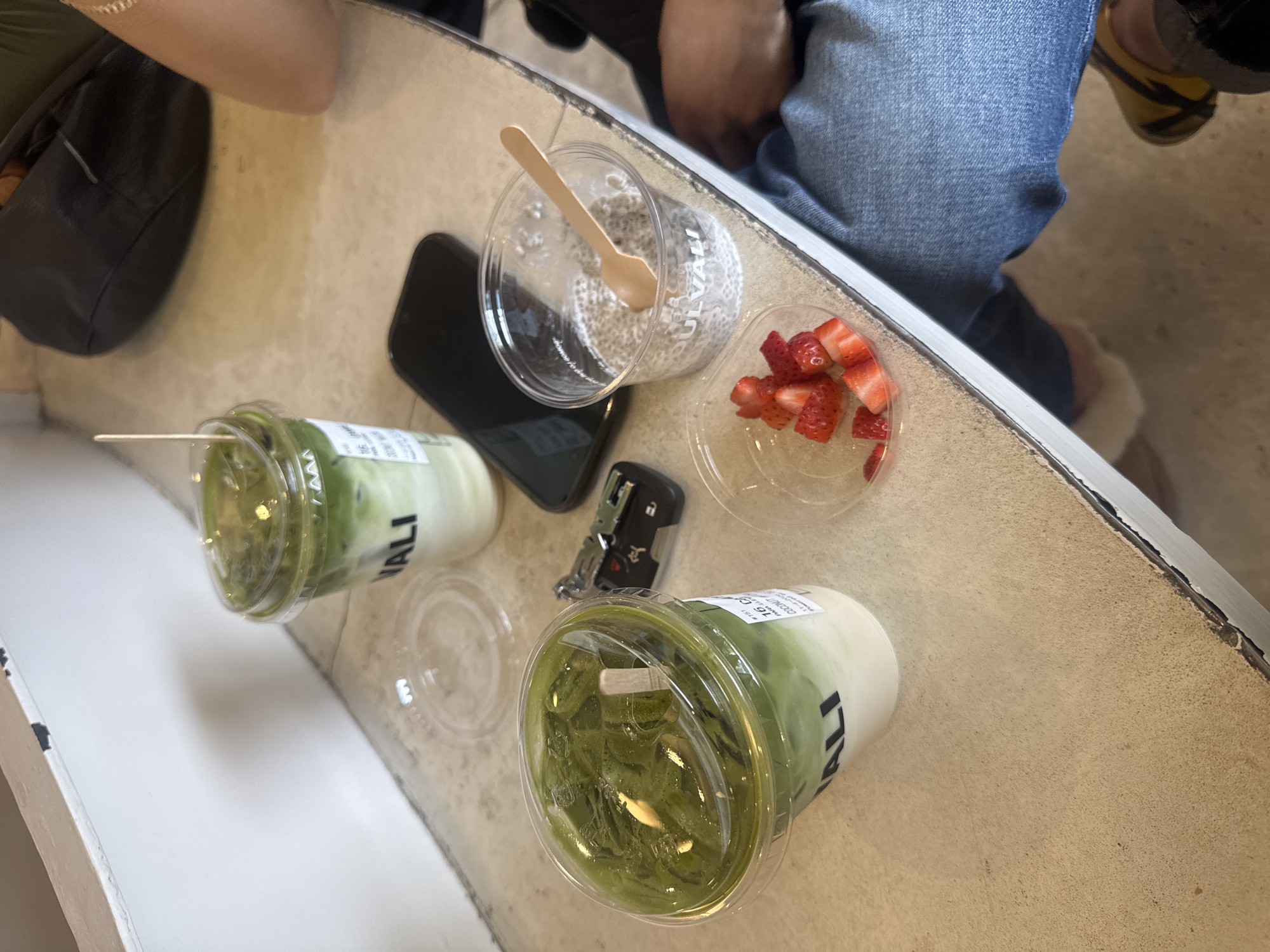 The image shows a tabletop scene with various items: two plastic cups filled with a green beverage labeled "VALI," a clear plastic cup containing a white, viscous substance with a wooden spoon inside, and a clear plastic container with sliced strawberries. A black smartphone and a car key fob are also on the table.
The table appears to be light-colored and has a slightly worn texture. In the background, a person wearing blue jeans and a light-colored shoe is partially visible, seated on the left side. Another person's black bag is seen on the left side. The location is likely a cafe or restaurant in Puebla, Mexico, due to the branding on the cups.