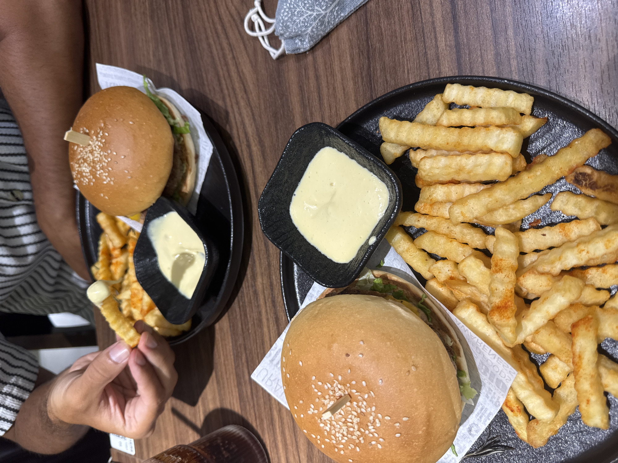 A wooden table is set with two black plates, each holding a hamburger, French fries, and a square black dish filled with a light-colored sauce. A person's hand is visible picking up a French fry and dipping it into the sauce. A glass of a dark liquid and a gray face mask are also present on the table. The location is likely Đà Nẵng, Vietnam.