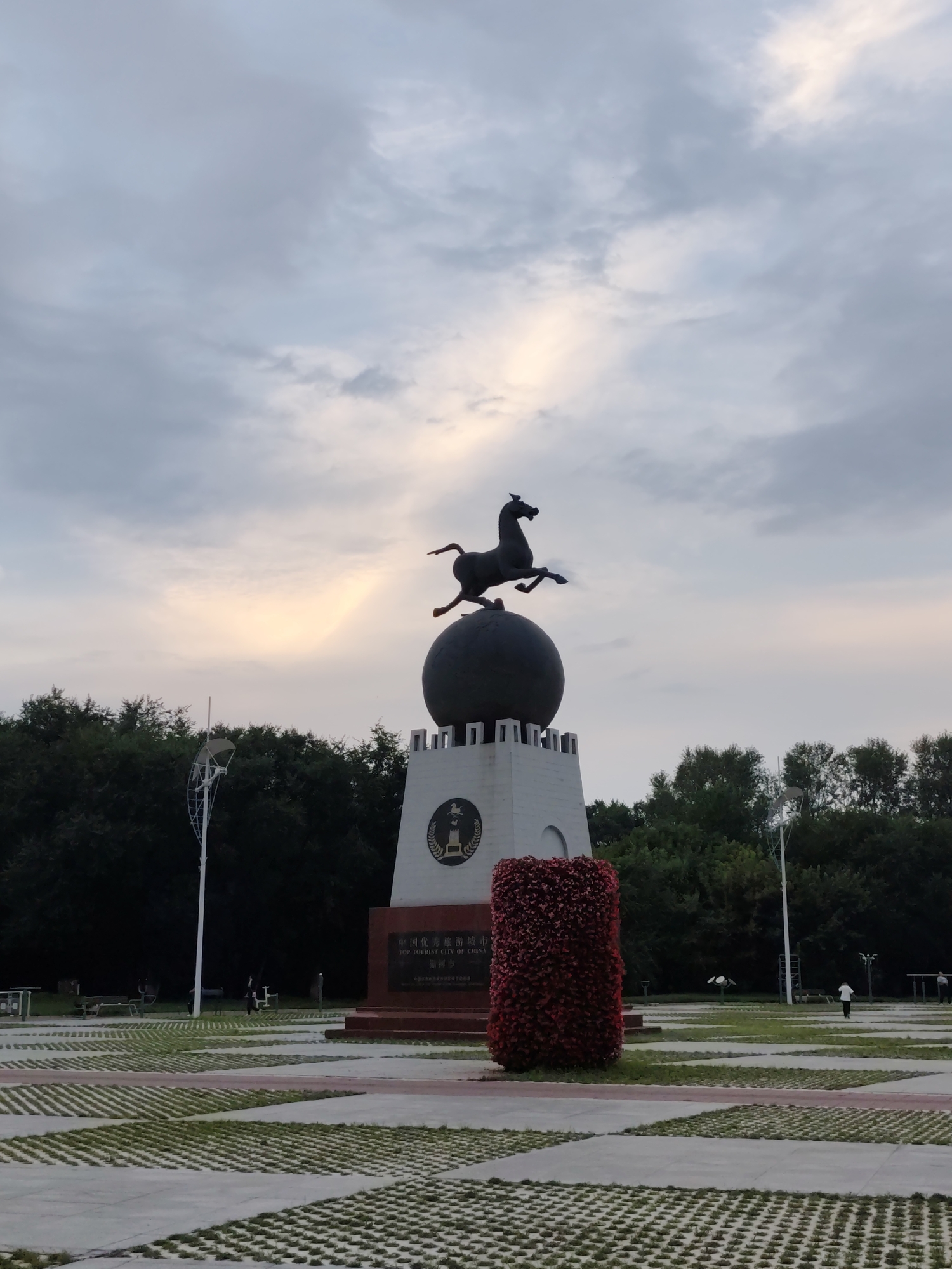 An outdoor, full-shot photograph, taken from a slightly low angle, shows a monument topped by a statue of a horse jumping over a globe. The monument is white and octagonal at the base, with a decorative band of red flowers. The globe is black, and the horse is a dark bronze. The monument is situated in a paved area with patches of grass. In the background, a line of trees forms a backdrop under a cloudy sky with hints of pink and yellow from a setting sun. Two lampposts with satellite dishes are visible to the left of the monument. In the distance, a few people are walking in the park. The overall scene suggests a public park or plaza.