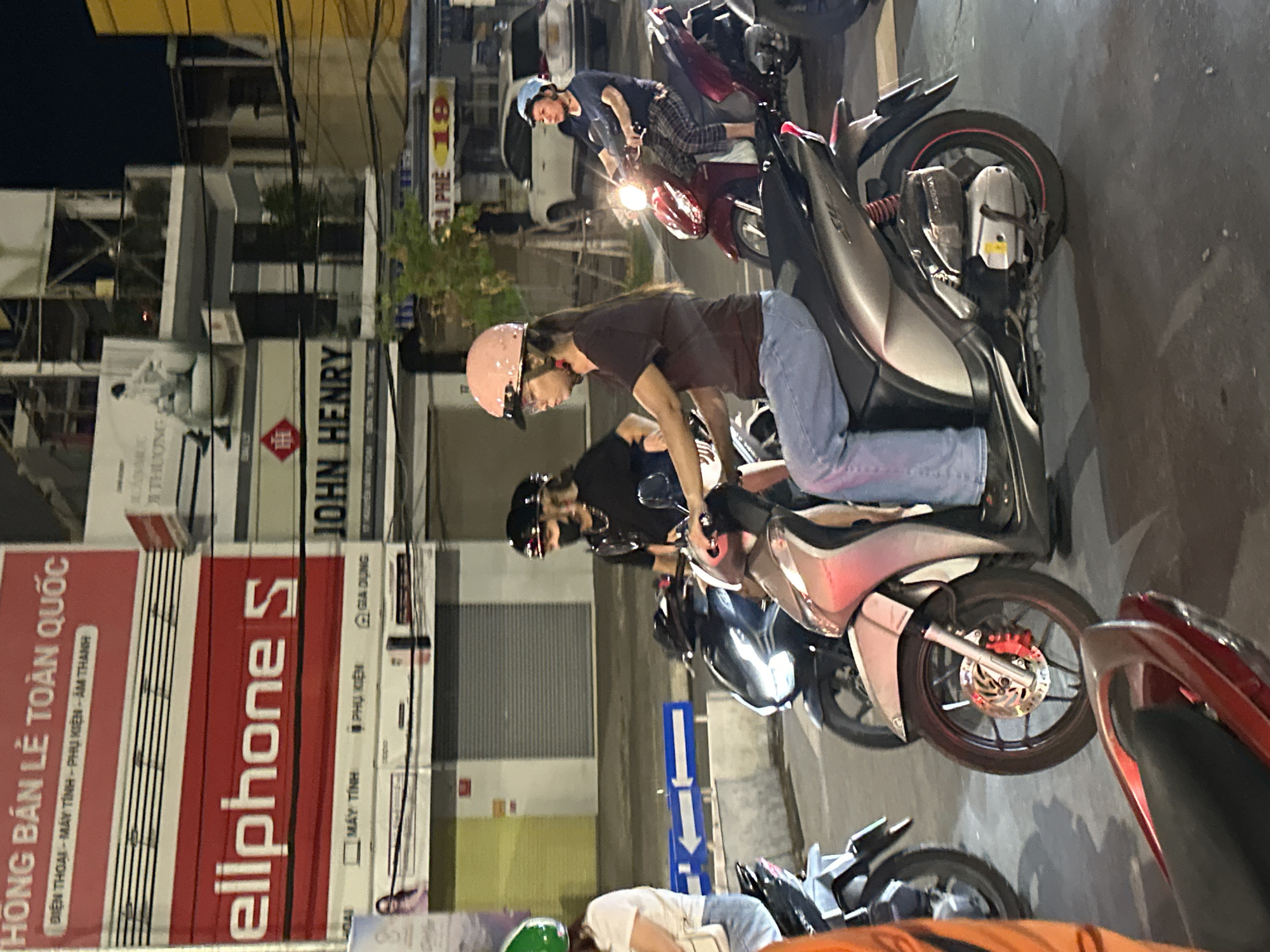 A nighttime street scene in Da Nang, Vietnam, features several people riding motorbikes. In the foreground, a woman wearing a pink helmet and light blue jeans rides a silver motorbike. To her left, another person wearing a green helmet rides a motorbike. Behind the woman in the pink helmet, two individuals are on a black motorbike, one wearing a dark helmet and the other a helmet with a face shield. Further back, a woman in a blue shirt and plaid pants rides a red motorbike. The background shows buildings with illuminated signs. One sign displays "HONGBANLE TOAN QUOC," and another reads "Ellphone S." The street is wet, indicating recent rain.