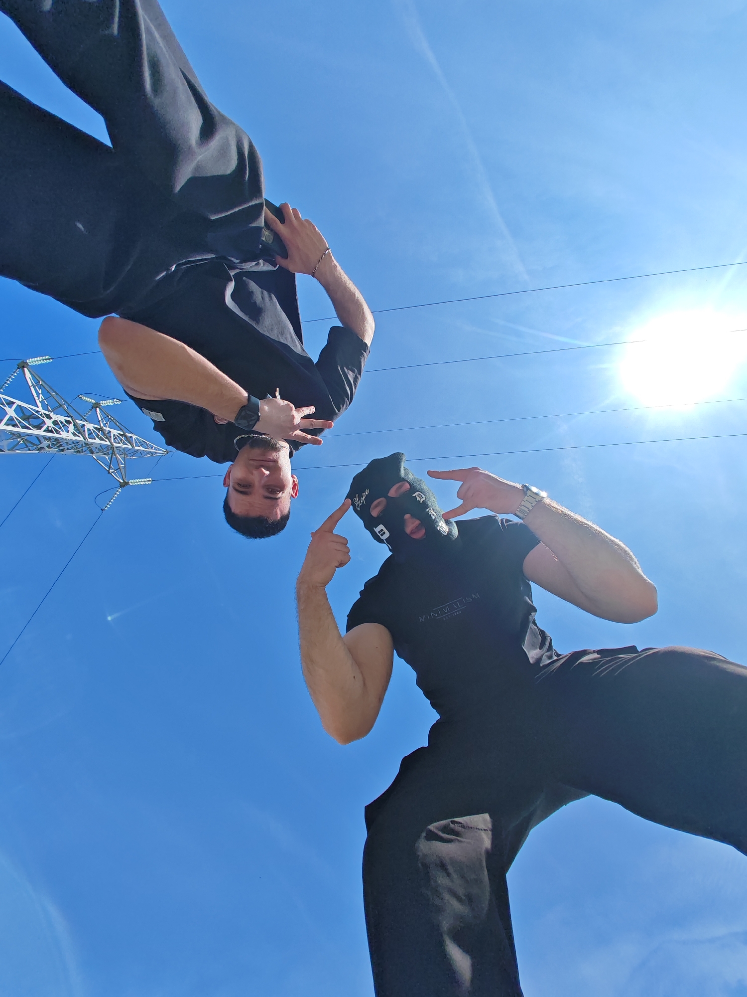 Two males are depicted in a low-angle photograph, positioned against a clear blue sky with a visible sun flare in the upper right quadrant. A large, lattice-structured electricity transmission tower is present in the upper left background, along with horizontal power lines.
The individual on the left appears inverted, wearing a black t-shirt and black pants. He has short dark hair and a short beard. His left wrist displays a dark watch, and his right hand is raised, forming a "rock-on" gesture. He is looking directly at the camera.
The individual on the right appears upright, wearing a black balaclava with white text or symbols, a black t-shirt bearing the text "MINIMALISM est. 1990," and black pants. A silver-toned watch is visible on his left wrist. His left hand is raised, making a "rock-on" gesture, while his right hand points towards the first individual.
The scene is illuminated by direct sunlight, casting minimal shadows and suggesting a bright day in Shumen, Bulgaria.