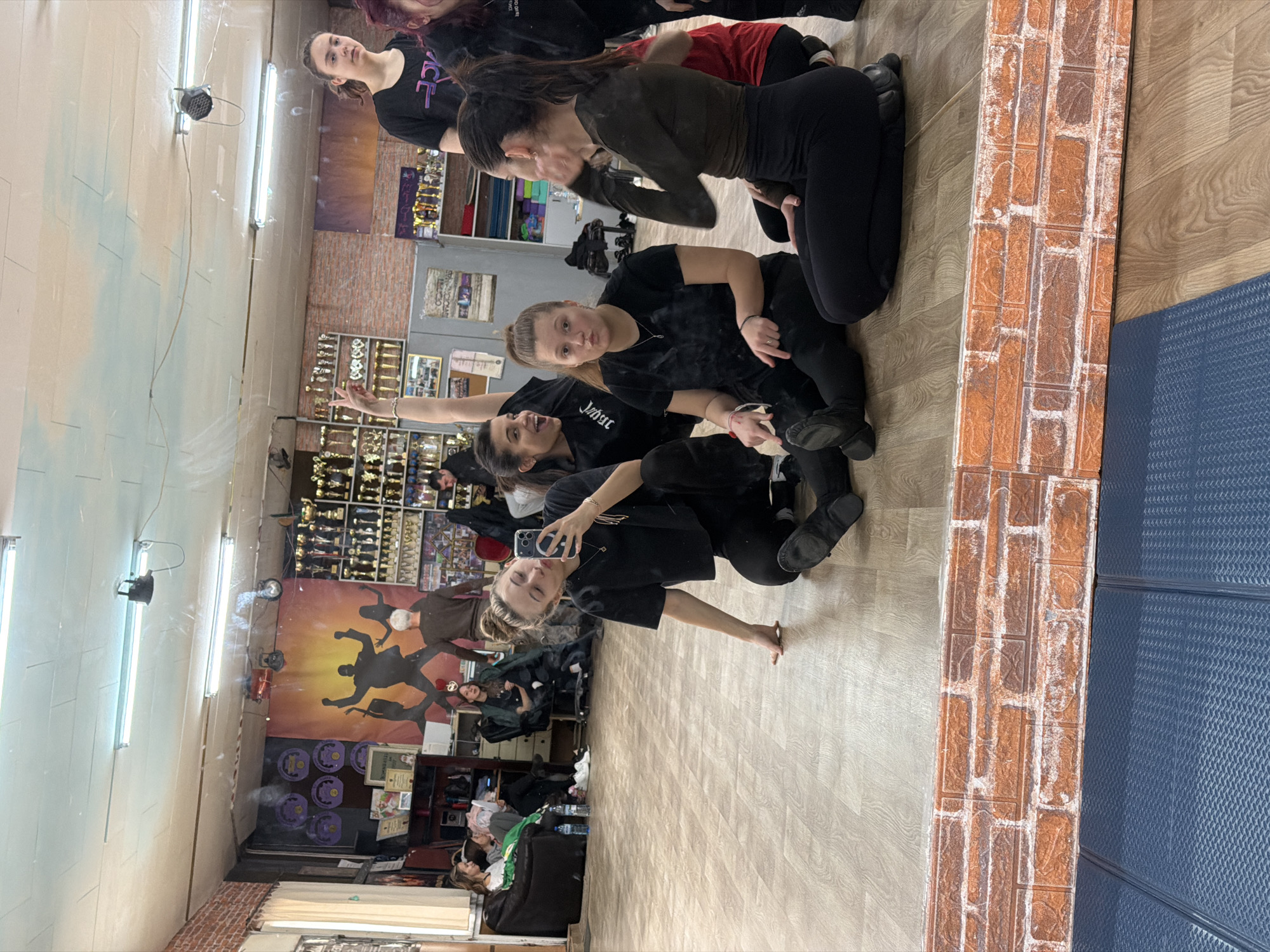 In a mirrored dance studio, a group of five young women are gathered close together on the floor, posing for a selfie. They are all dressed in black clothing, with the exception of one woman wearing a brown top and another wearing a red top. The woman taking the photo is holding a smartphone and smiling at the camera. The reflection reveals the studio's interior: awards and trophies on shelves, a mural depicting dancers, and seating area. The studio appears to be located in Plovdiv, Bulgaria.