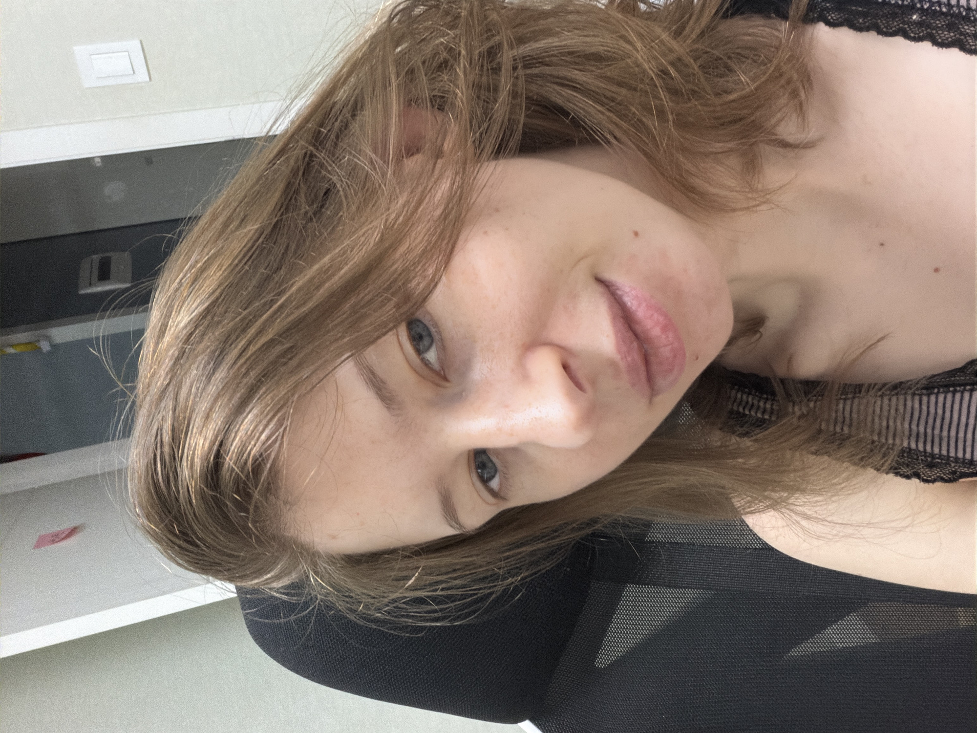 A close-up, selfie-style portrait of a fair-skinned young woman with light brown hair and blue eyes is captured in what appears to be an office environment in Dongducheon-si, South Korea. She is seated in a black mesh-backed office chair, her head tilted slightly to her left. The background is softly blurred, showing a white wall with a light switch and a red sticky note. To the left of the woman, a portion of the black office chair's headrest is visible. Her hair is loose and slightly tousled, falling around her face and shoulders. She is wearing a black top with thin stripes and lace trim. The lighting is bright, suggesting an indoor setting.