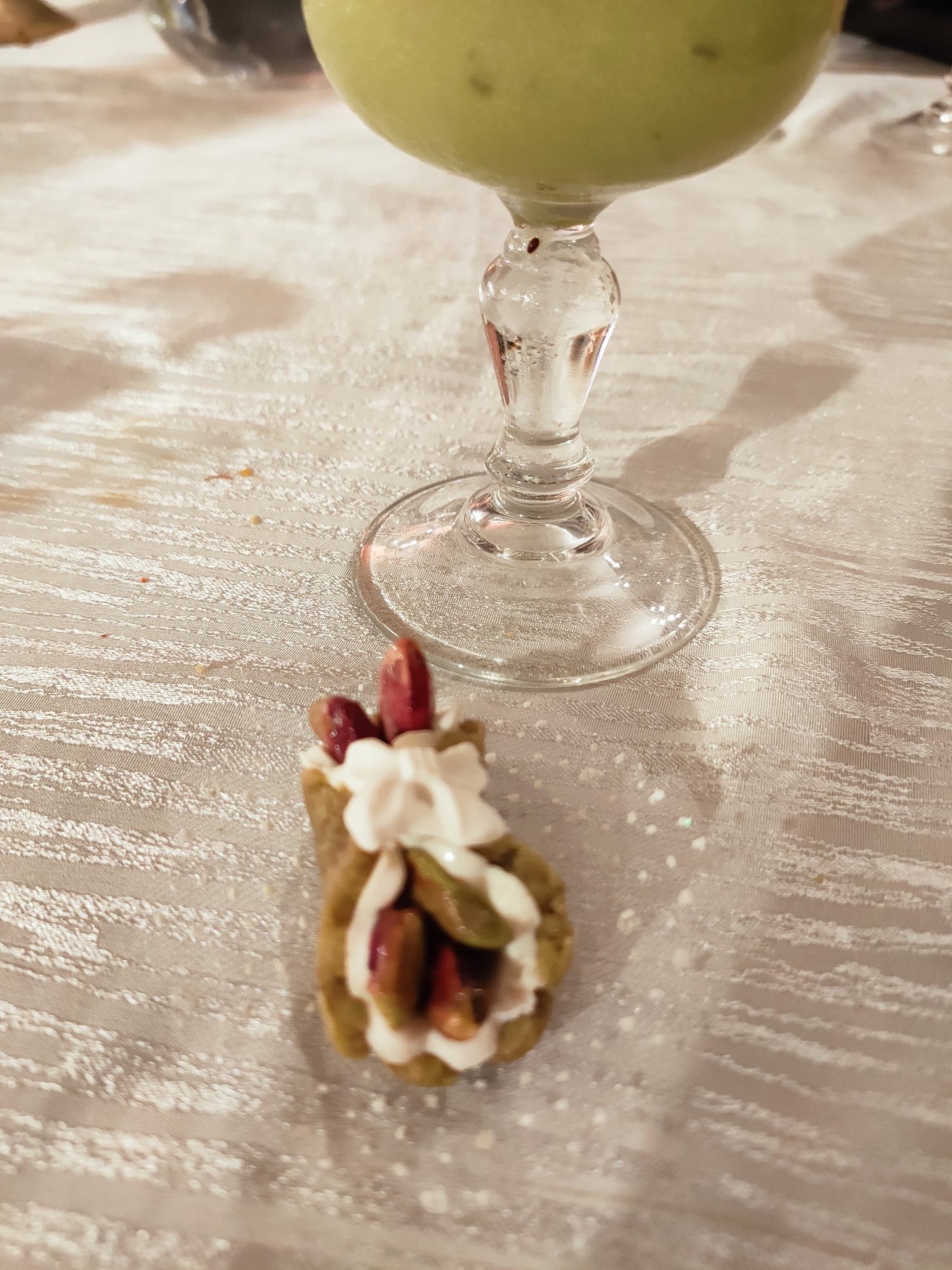 A close-up, slightly overhead view of a single cannoli and a glass of green liquid on a textured, light-colored tablecloth. The cannoli, positioned in the lower center of the frame, is filled with white cream and appears to have pieces of red fruit and green nuts visible. The glass, which has a clear, stemmed base, contains a pale green, frothy beverage and is located in the upper right quadrant of the image. The tablecloth has a raised, woven pattern with some scattered white powder, possibly sugar or flour, on its surface. The lighting creates soft shadows cast by the glass and the cannoli. The image does not contain any information to indicate a specific location in Mornag, Tunisia.