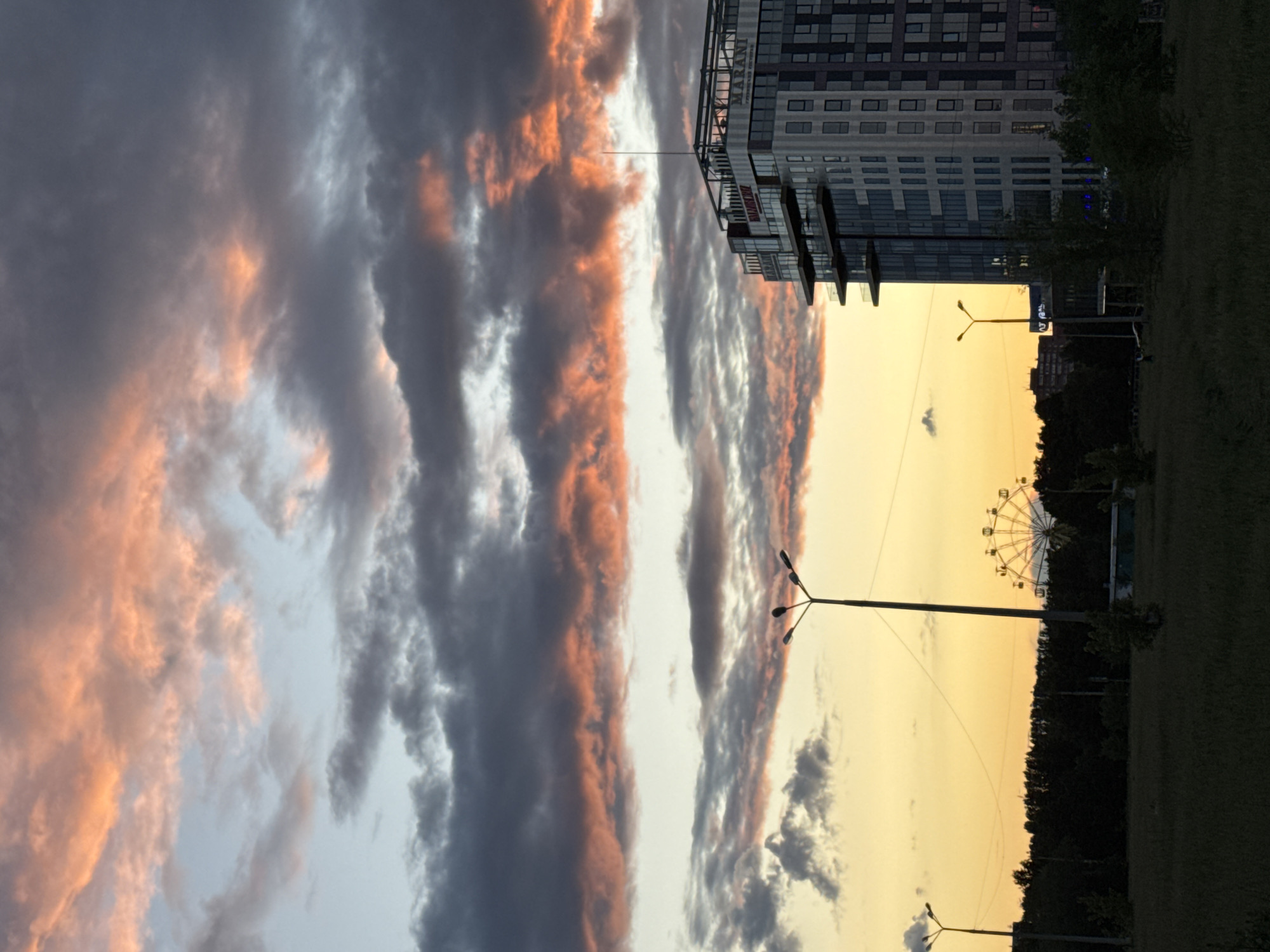 The image depicts an outdoor scene in what appears to be a city, possibly in Unknown, Unknown. A Ferris wheel is visible in the distance against a yellow-orange sky, suggesting either sunrise or sunset. Several streetlights are present. Adjacent to the sky is a modern building with multiple floors and a glass facade. The sky is filled with dramatic clouds, some tinted with orange hues. Trees and grass can be seen in the foreground.