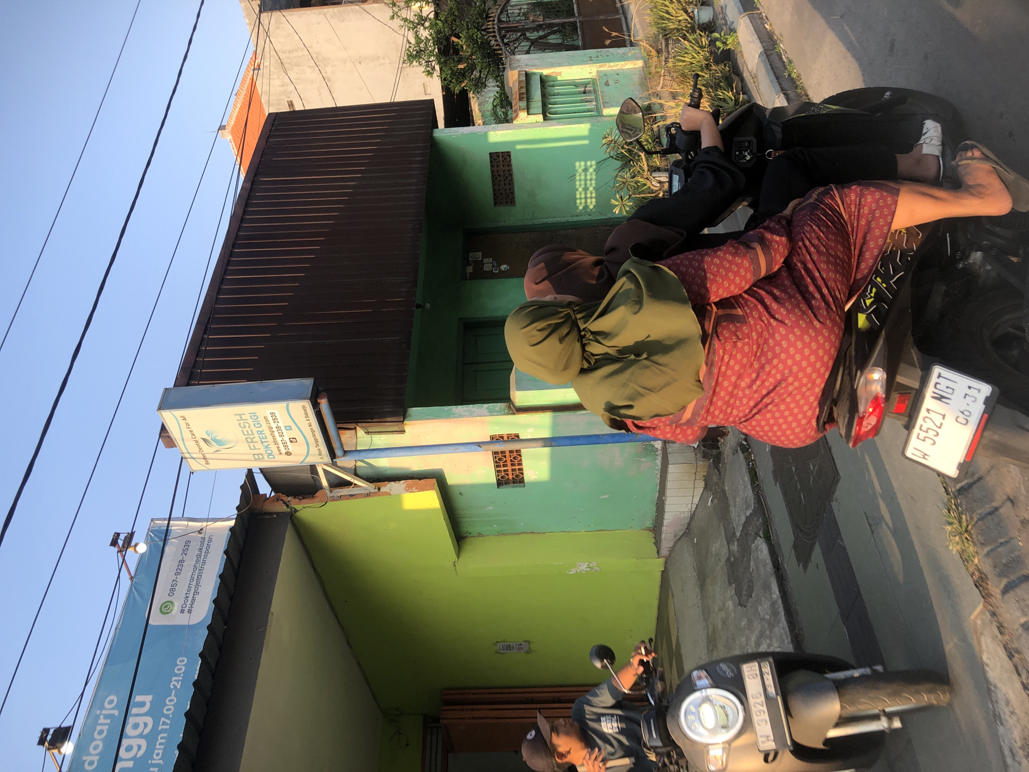 The image depicts a street scene in Boro, Indonesia, under a clear blue sky with visible overhead wires. In the foreground, two people are riding on a dark-colored scooter. The person in the back is wearing a green hijab and a patterned red dress. The person in front, partially visible, is wearing a black shirt and a black cap. The scooter has a license plate that reads "W 5521 NGT 06-31". To the left, another scooter is parked, with a license plate "H 3926 BH". A man wearing a dark t-shirt and a black baseball cap is sitting on this scooter, looking towards the left.
The buildings in the background are painted in vibrant green and light green hues. A sign on a light blue background is visible above the green building, with text that includes "0857-9238-2539". Another sign, white with blue lettering, is mounted on a pole and reads "B FRESH DOKTER GIGI" and provides a website and phone number. The street is paved, and there is some vegetation visible on the right side of the image, including bushes and small trees. The lighting suggests it is either early morning or late afternoon, with sunlight casting shadows on the street and buildings.