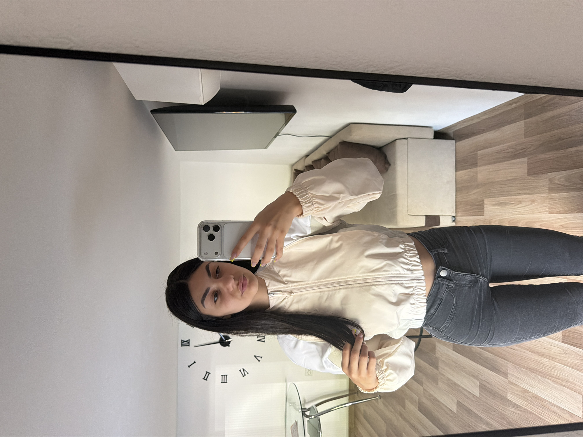 A young woman takes a selfie in a mirror. She is wearing gray jeans and a light-colored jacket with white accents. A clock is visible on the wall behind her. In the background, there is a television, couch, and a table. The flooring is wood-patterned. This scene appears to be taking place in an apartment located in Gabrovo, Bulgaria.