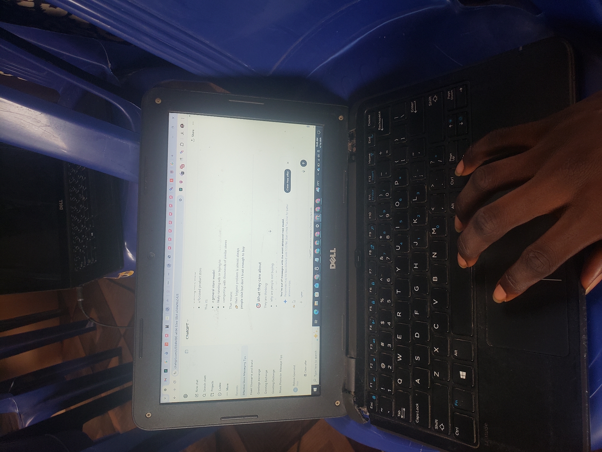 A person with dark skin is typing on a black Dell laptop. The laptop is open, displaying a webpage with the ChatGPT interface. The screen shows recent conversations and some suggestions, along with a "Claim free offer" banner. The laptop is placed on a blue plastic chair. A second black laptop, also a Dell, is visible in the background, slightly out of focus. The location appears to be indoors. There is no indication this image was taken in Alalubosa, Nigeria.