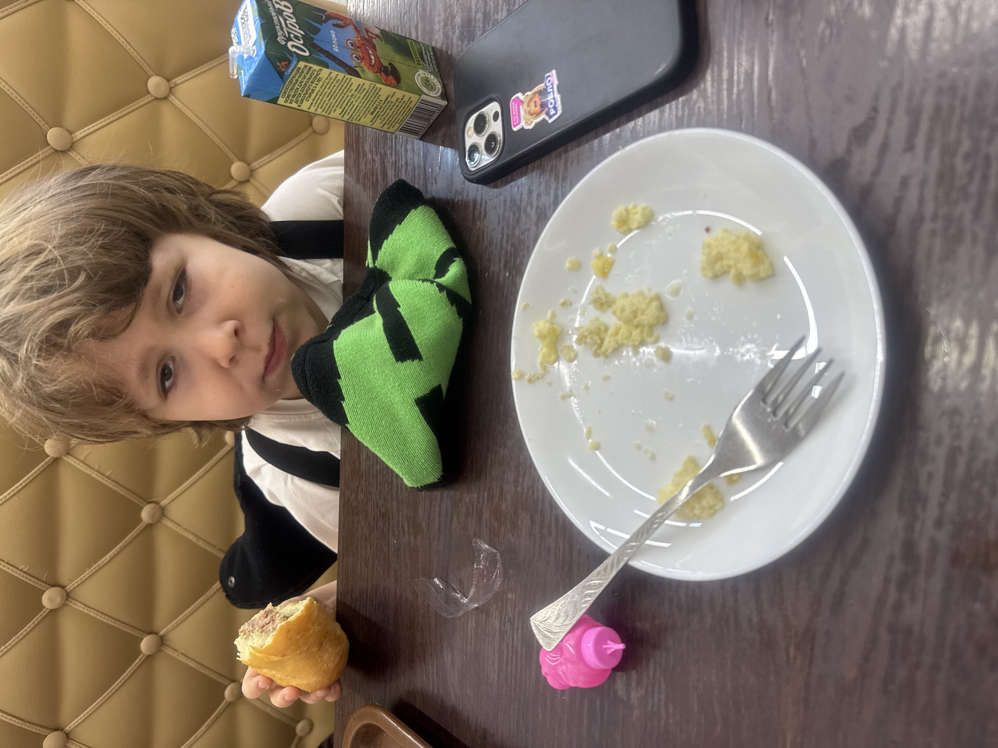 The image shows a child at a table with food and other items. The child is holding a pastry in their left hand and looking at the camera. On the table, there is a plate with crumbs, a fork, a small pink toy, a juice box labeled "Остров" (Ostrov), and a cell phone with a sticker. The background includes a tufted wall. The juice box suggests the location might be in Russia, and the OCR of the juice box is showing "Яблоко" (Apple) which supports this notion. The juice box also indicates "ДЕТЕЙ ДОШКО" for children. The combination of the juice box and the child suggests the picture was taken in Chaikovsky, Russia.