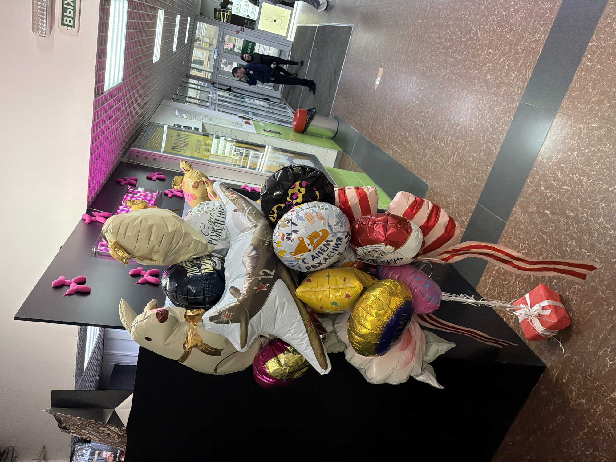 The image depicts a bouquet of various helium-filled balloons, along with a small gift wrapped in red paper with a white ribbon, placed on a black surface.
The balloons include round balloons with printed designs, as well as shaped balloons, such as a star, a unicorn, and a zebra. One of the round balloons features a design with the phrase "С Днем Рождения" (Happy Birthday). Red and white striped ribbons are tied to the balloons and the gift.
The bouquet is situated in a commercial setting, possibly a shopping mall or store. In the background, there is a corridor leading to an entrance/exit area with people walking through the doors. The floor is tiled in a pattern of brown and beige. There are visible shop signs and a display case in the background. The location appears to be Birobidzhan, Russia, based on the Cyrillic text on the balloons.
