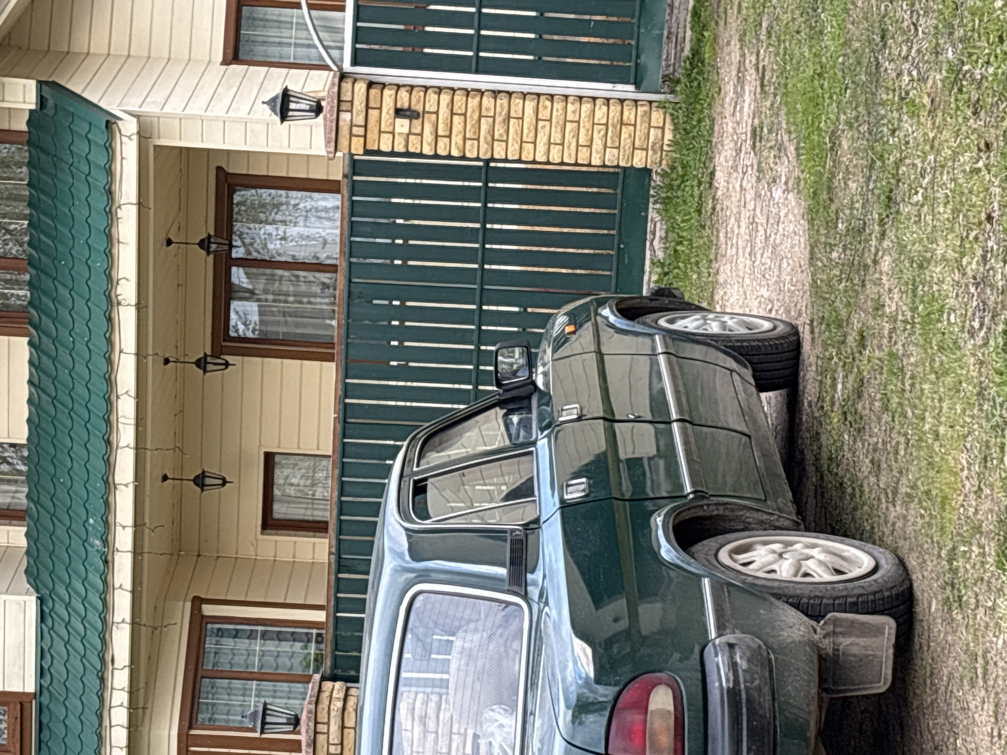A medium shot depicts a dark green car parked adjacent to a yellow-sided building with a green tiled roof, and a green wooden fence. The car appears to be an older model. The setting is outdoors, next to a grassy area and a dirt path. Black lantern-style lamps hang from the building's eaves. The image's original location is Zaprudnya, Russia.