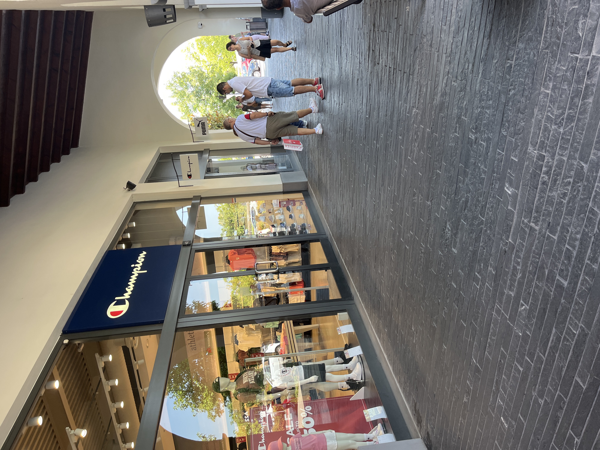 The image shows the exterior of a Champion store in an outdoor mall setting, possibly an outlet mall, as suggested by the presence of multiple brand stores and the architectural style. The Champion store has a large glass storefront displaying mannequins dressed in athletic wear and sale signs. Next to the Champion store is another branded store, possibly Puma, indicated by a hanging sign. Several people are visible walking along the covered walkway in front of the stores, some carrying shopping bags. The walkway is paved with dark, rectangular stones. The architecture includes arched openings and a wooden-beamed ceiling. The scene appears to be in Rodengo, Italy, as indicated by the context provided. The lighting suggests it is daytime, with sunlight filtering through the trees and the open areas of the mall.