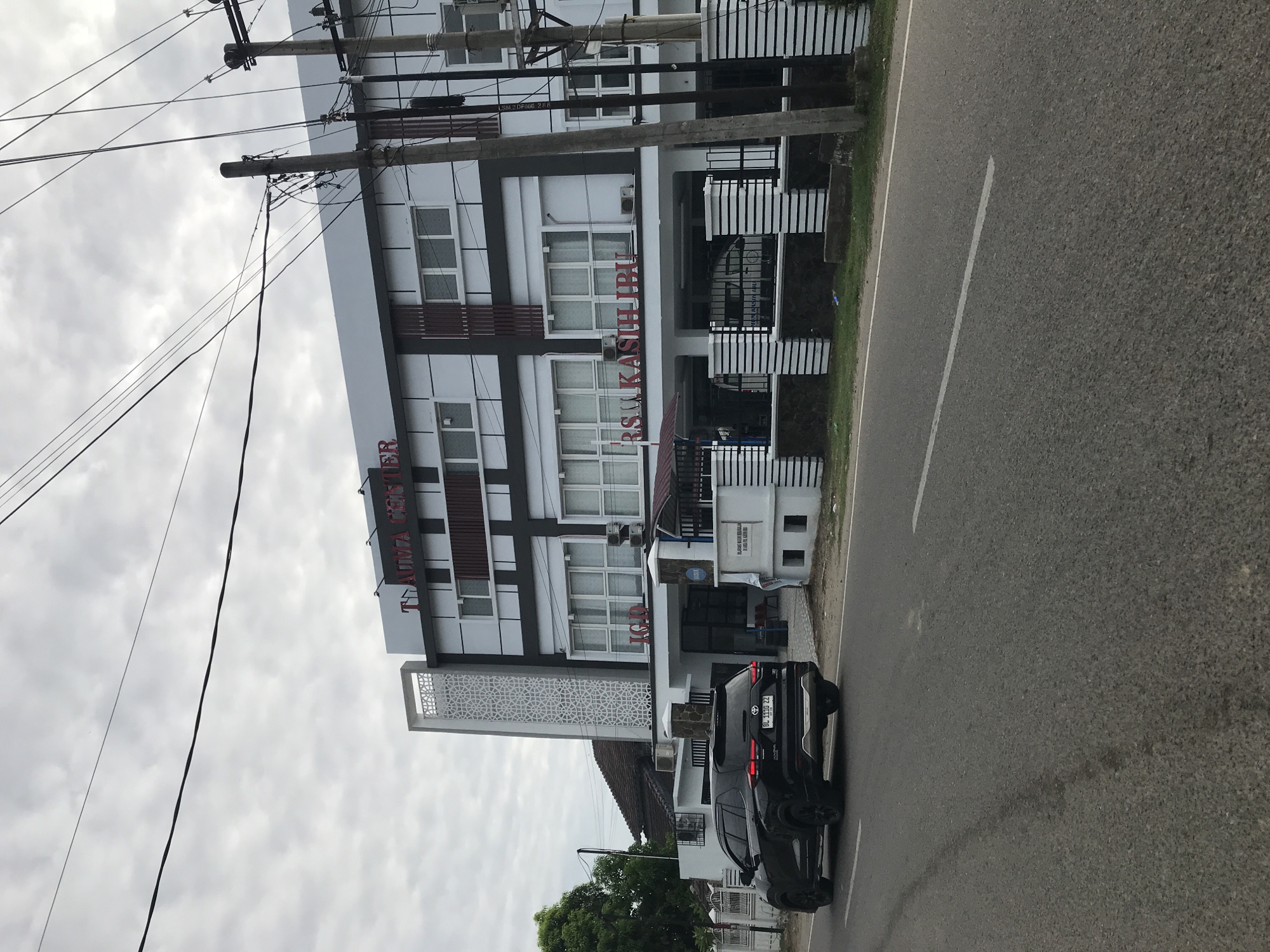 The image shows the building of the "T. Aima Center - RSU Kasih Ibu" hospital in Lhokseumawe, Indonesia, as indicated by the text on the building. The modern, multi-story building is white with gray and dark brown accents. The entrance area has a gated section. Two dark-colored cars are parked in front of the building, along the side of a two-lane asphalt road with a painted white line. Power lines and utility poles are visible on the left side of the frame. The sky is overcast.